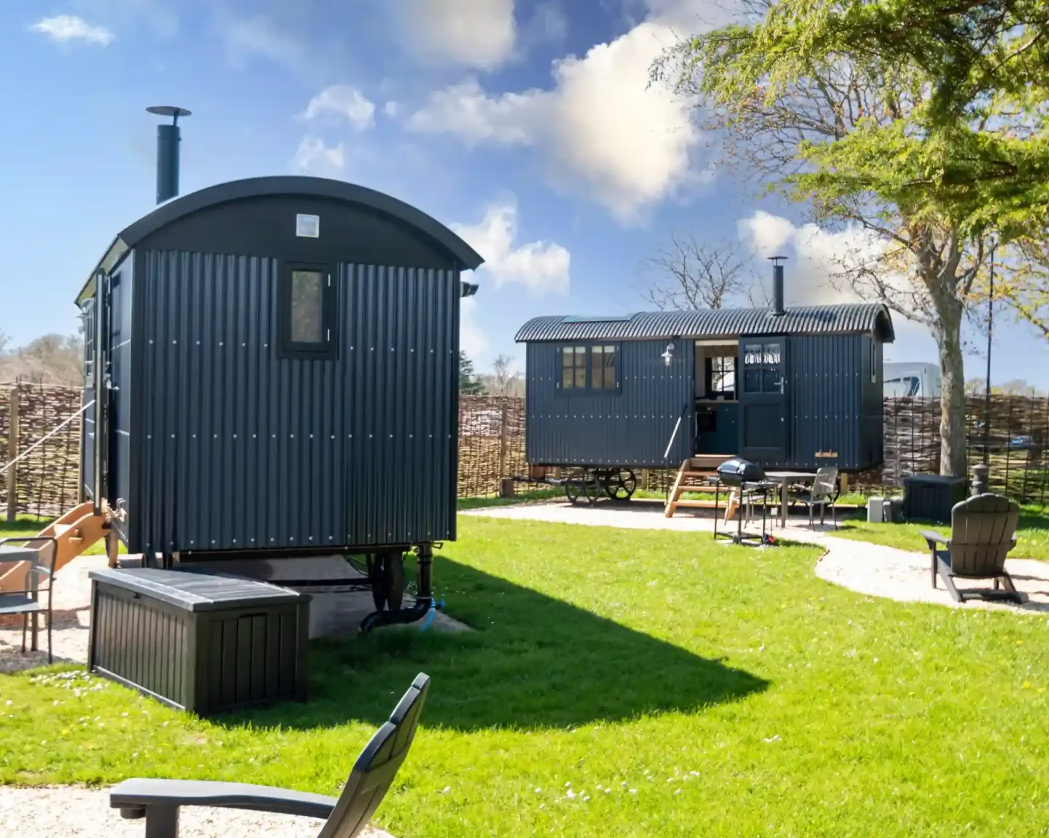 Two modern shepherd huts with dark corrugated exteriors set on a grassy lawn with outdoor seating, a barbecue grill, and a tree under a blue sky with scattered clouds.