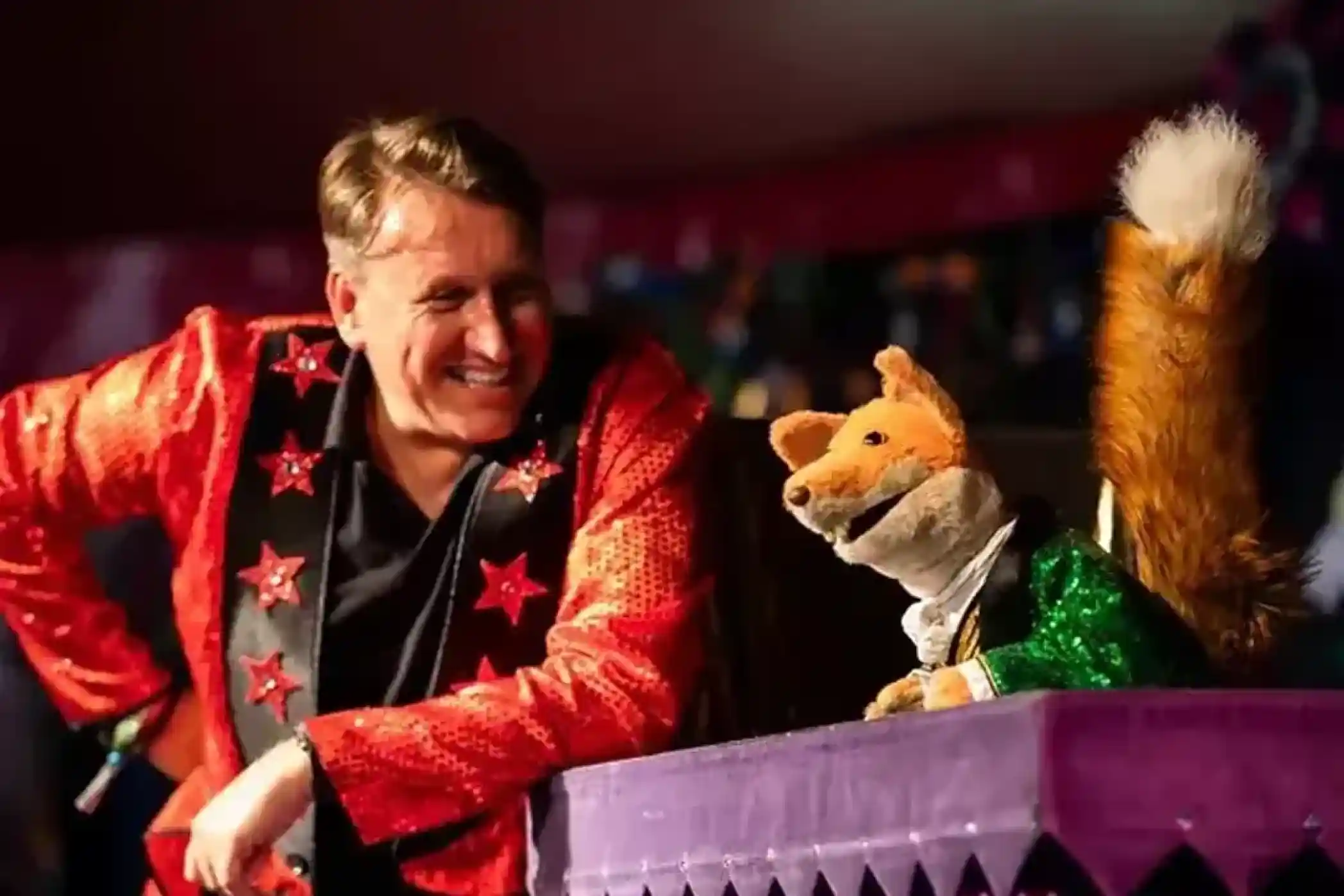 A performer in a sparkling red and black jacket smiles at a animated puppet fox wearing a green jacket, both engaging in a playful interaction on stage.