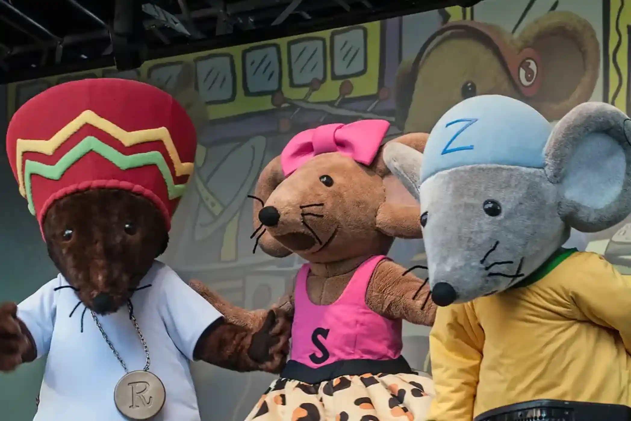 Three costumed mouse characters perform on stage. The first mouse, wearing a red and yellow hat, has a medal around its neck. The second mouse, dressed in a pink top with a black skirt, has a pink bow. The third mouse, wearing a blue cap and green shirt, is positioned next to them. A colourful backdrop is visible behind them.