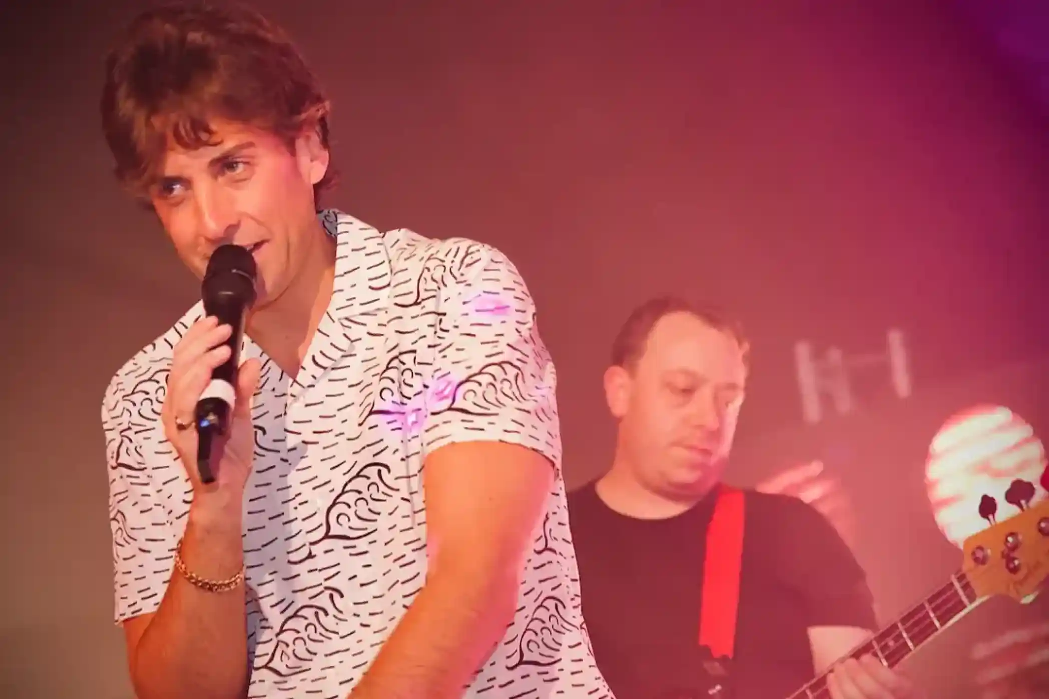 A male singer with curly hair holds a microphone while performing on stage, wearing a patterned short-sleeve shirt. In the background, a guitarist plays with a focused expression, illuminated by stage lighting.