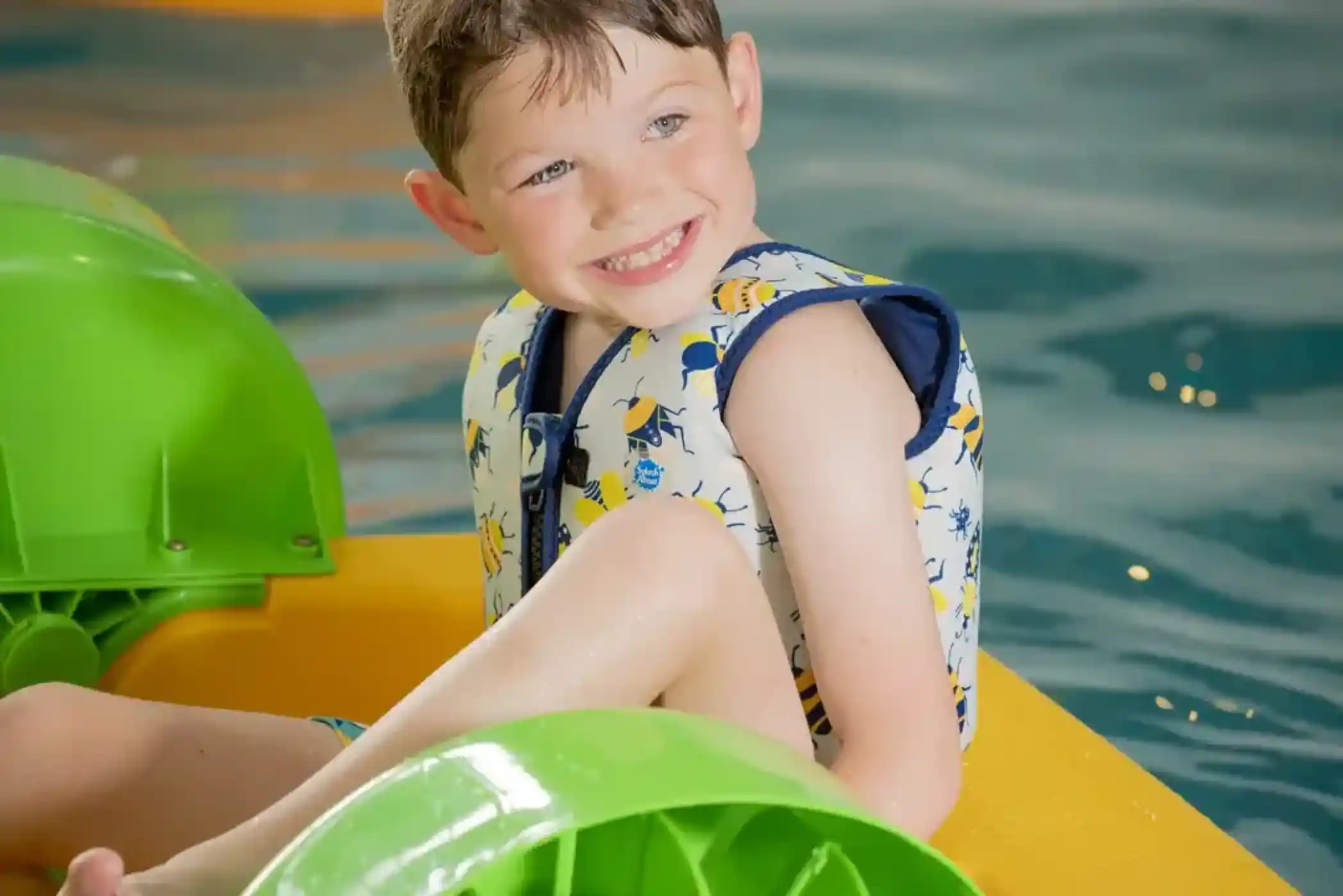A smiling boy wearing a patterned life jacket sits in a colourful paddle boat on water, with bright green paddles visible.