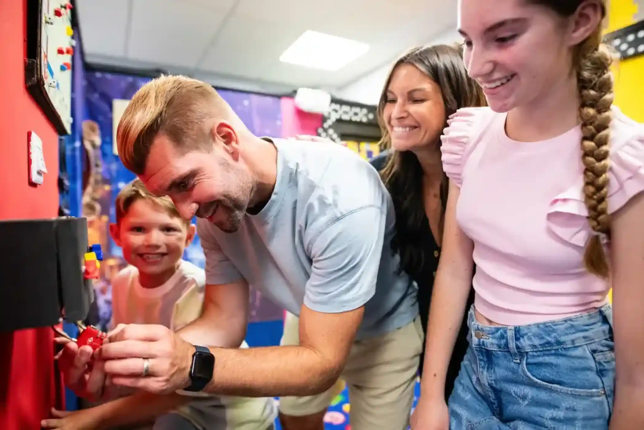 A smiling family works together on a puzzle in a colourful escape room. A man adjusts a lock on the wall while a young boy, woman, and teenage girl watch and laugh, enjoying the interactive challenge.
