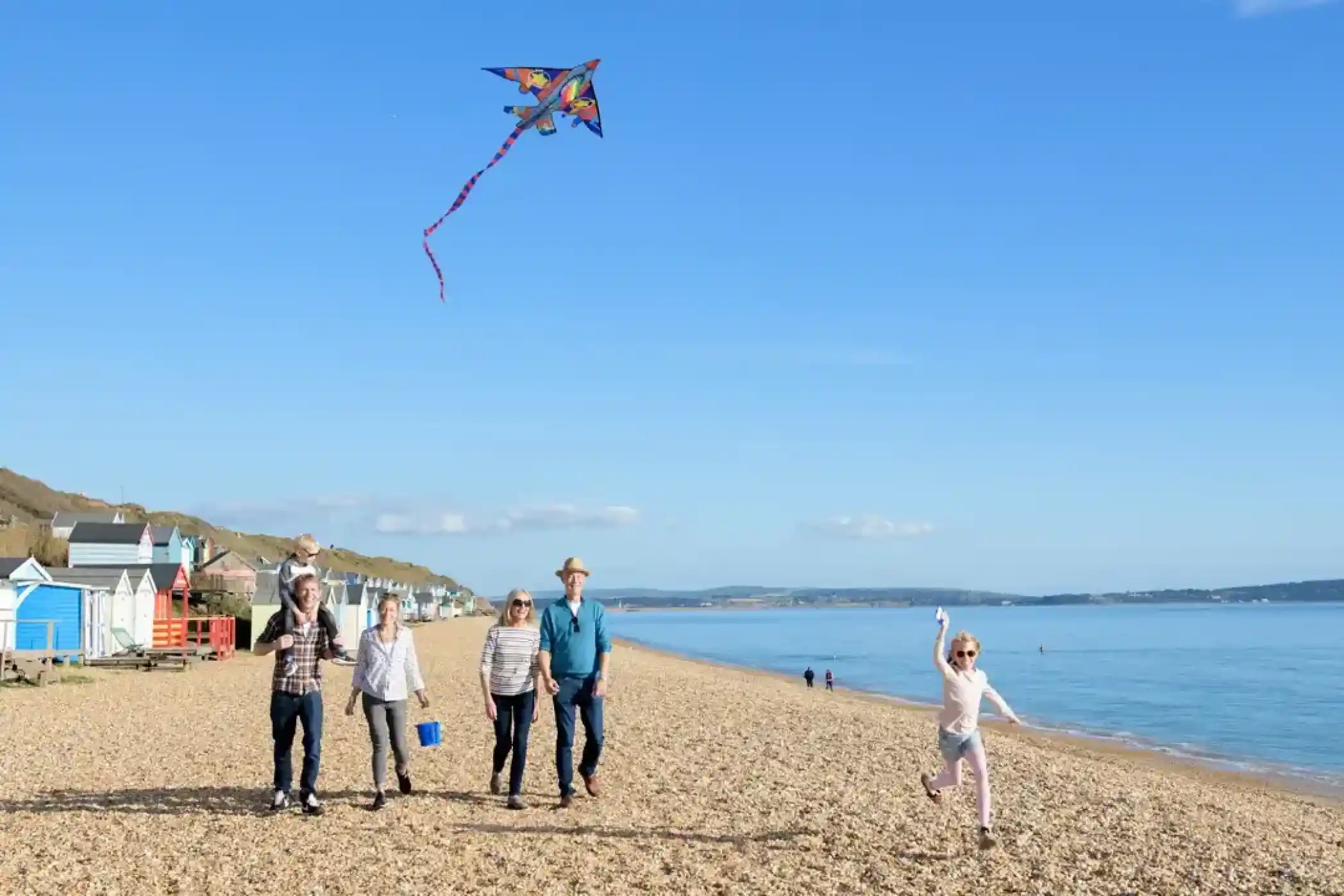 Family enjoys a sunny day flying a kite on a pebble beach with colourful beach huts in the background.
