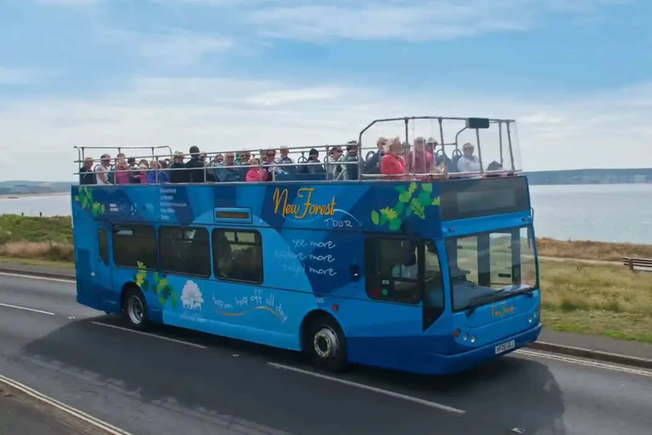 A blue double-decker New Forest Tour bus with passengers on the top deck drives along a coastal road.
