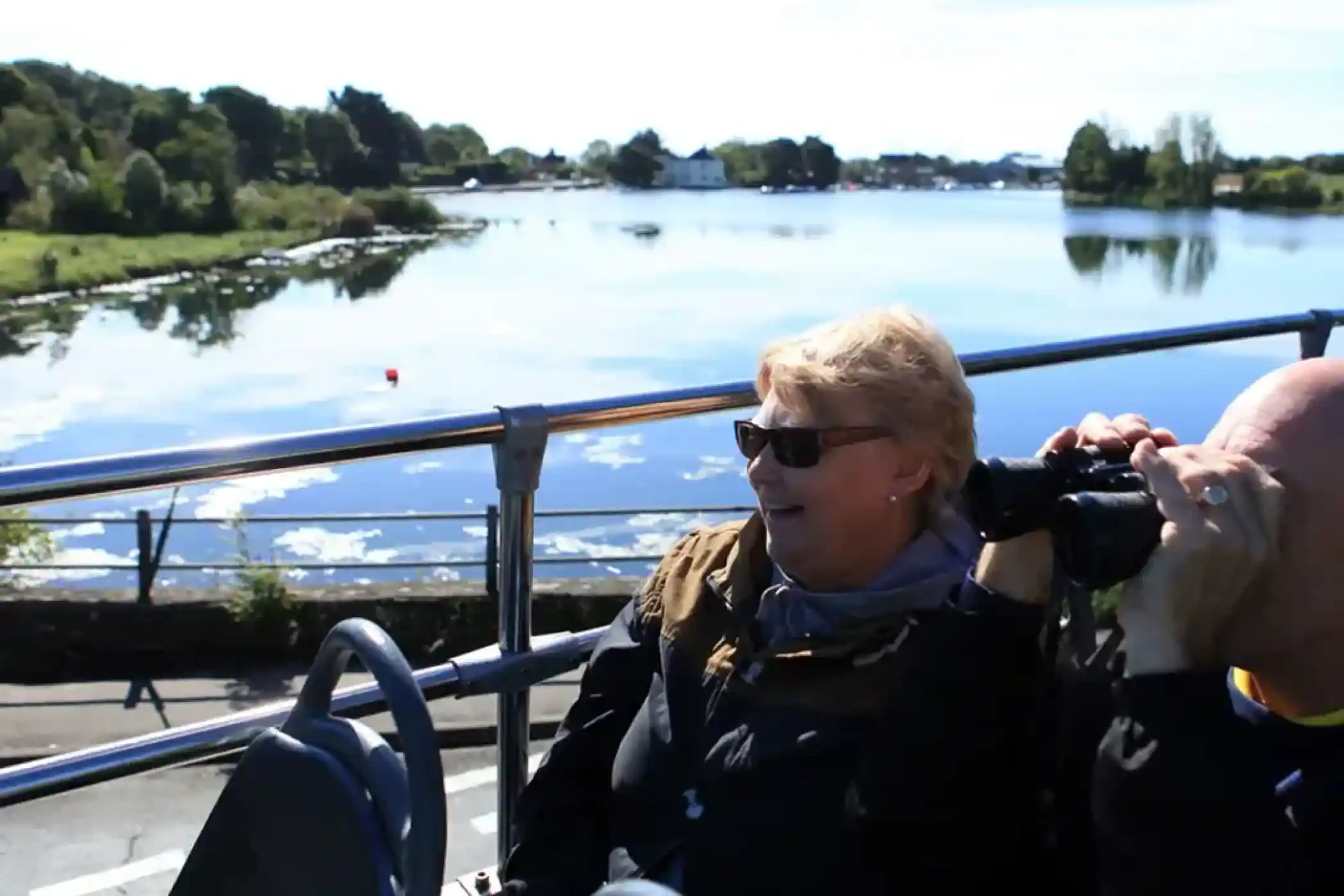 Two people on an upper deck of a bus look out at a lake with trees and buildings on the far shore.