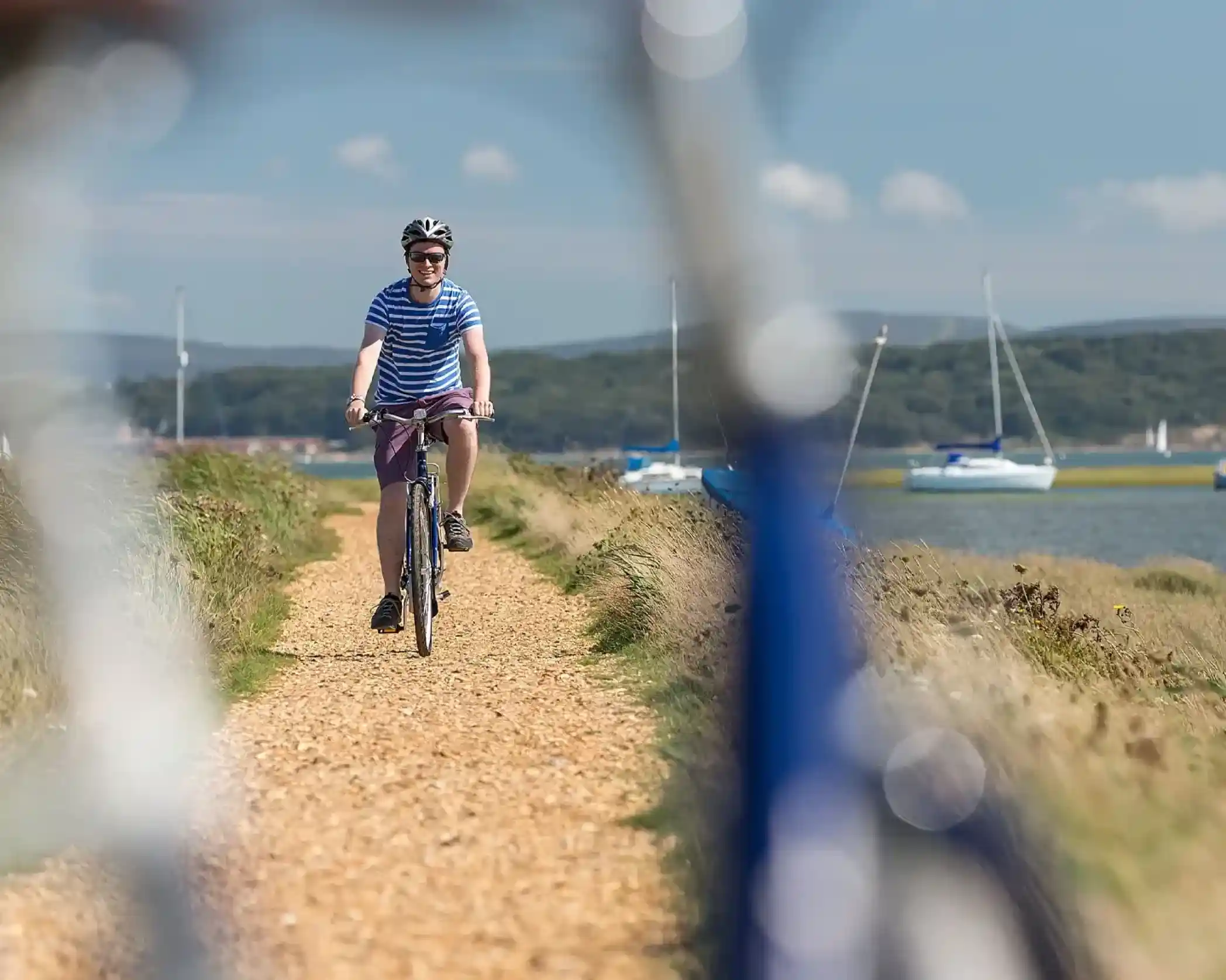 Man in striped shirt rides a bicycle on a gravel path with sailboats in the background.