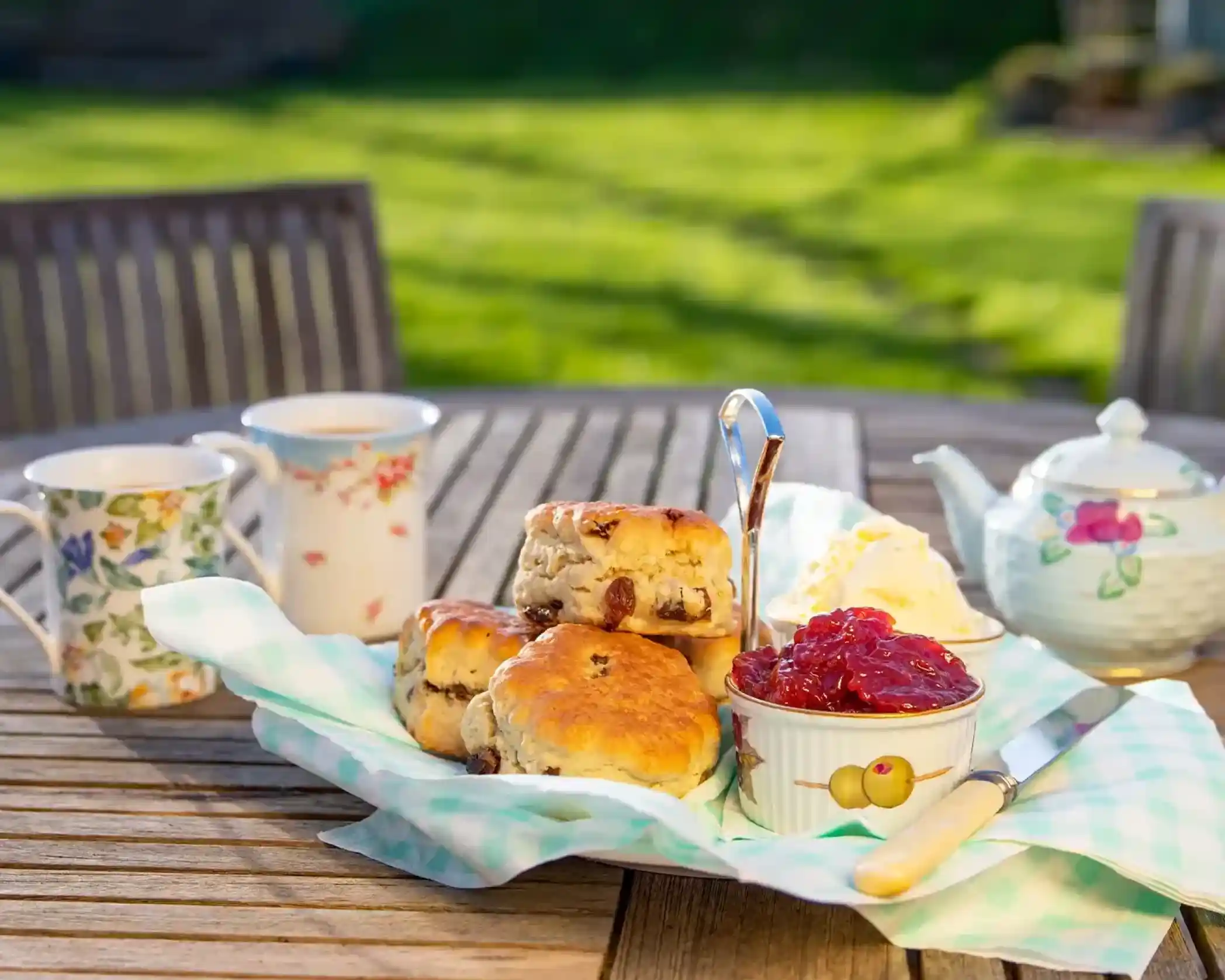 A tiered stand holds scones, clotted cream, and jam, with two floral teacups and a teapot on a wooden table in a garden.