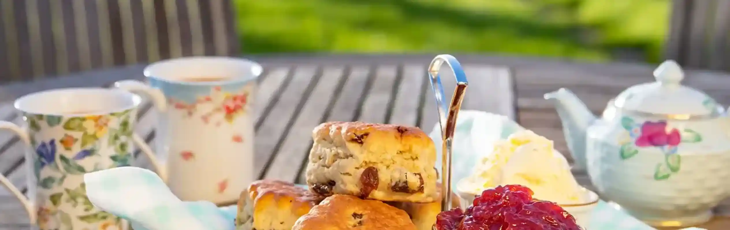 A tiered stand holds scones, clotted cream, and jam, with two floral teacups and a teapot on a wooden table in a garden.