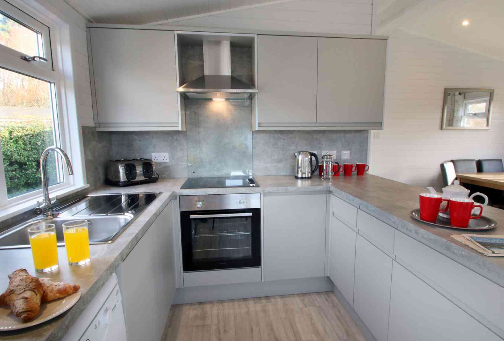 A modern kitchen with gray cabinets and a stainless steel oven. There are two glasses of orange juice and a croissant on the counter, along with a kettle and several red mugs. A dining table can be seen in the background.