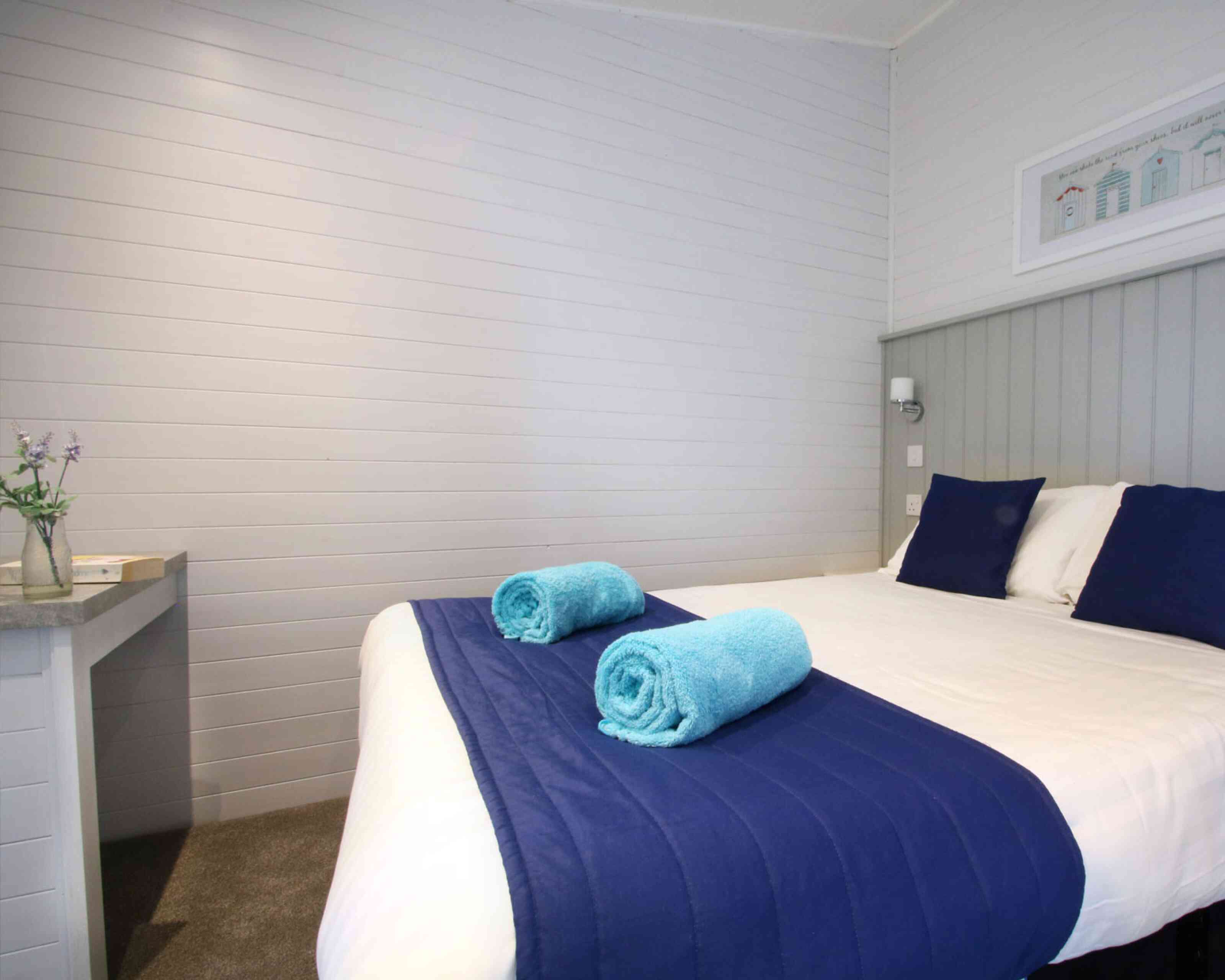 A cozy bedroom featuring a neatly made bed with white linens, a blue blanket, and two rolled turquoise towels. A small table is positioned against the wall, and there are light gray wooden walls with a decorative frame on display.