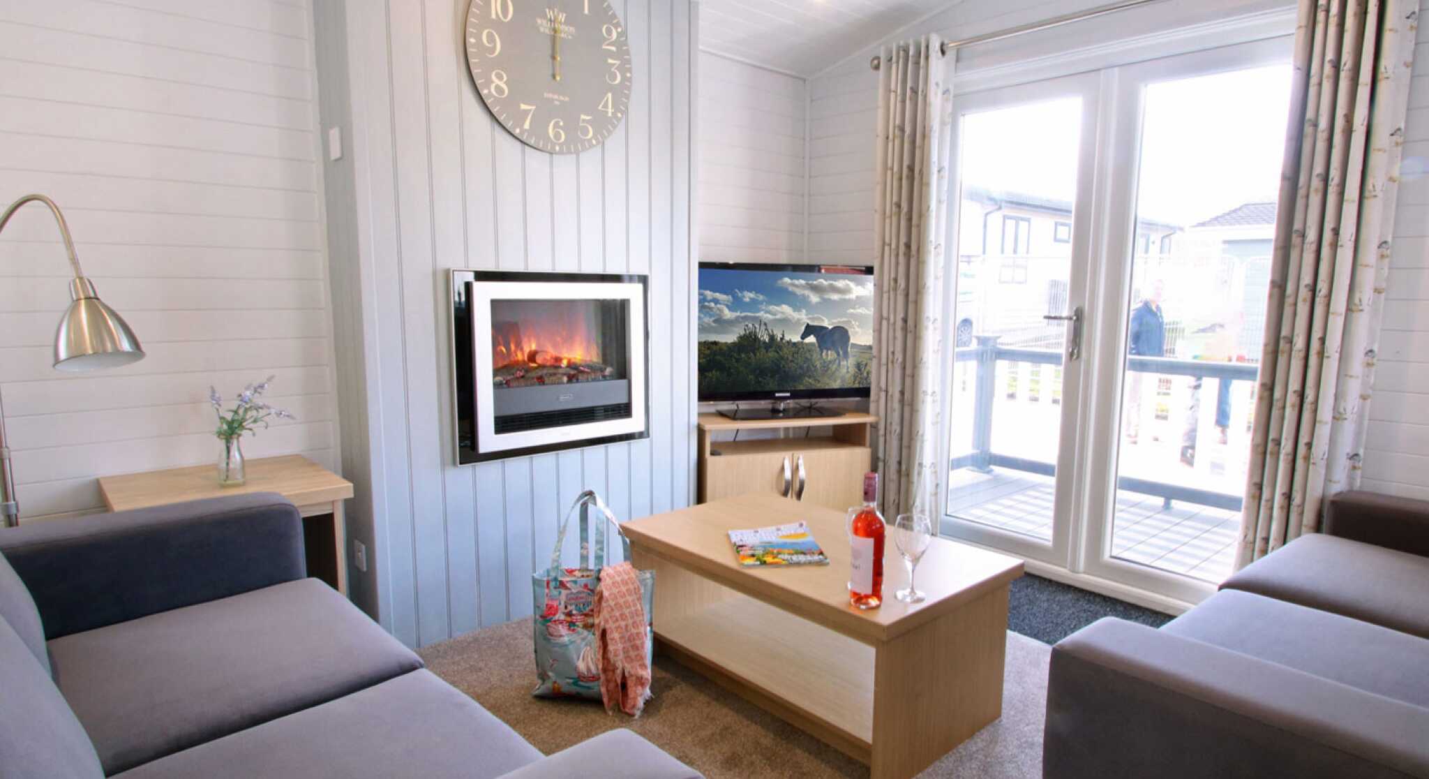 A cozy living room featuring two gray sofas, a coffee table with magazines and a drink, and a wall-mounted electric fireplace. A large clock hangs on the wall, and sliding glass doors open to a deck with outdoor views.