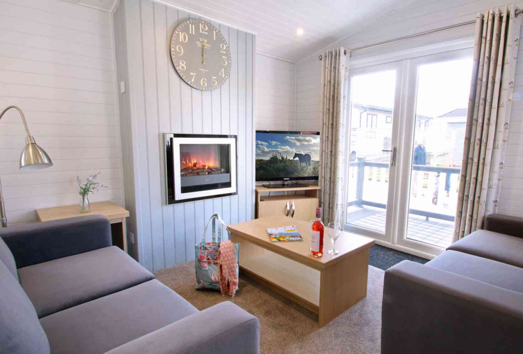 A cozy living room featuring two gray sofas, a coffee table with magazines and a drink, and a wall-mounted electric fireplace. A large clock hangs on the wall, and sliding glass doors open to a deck with outdoor views.