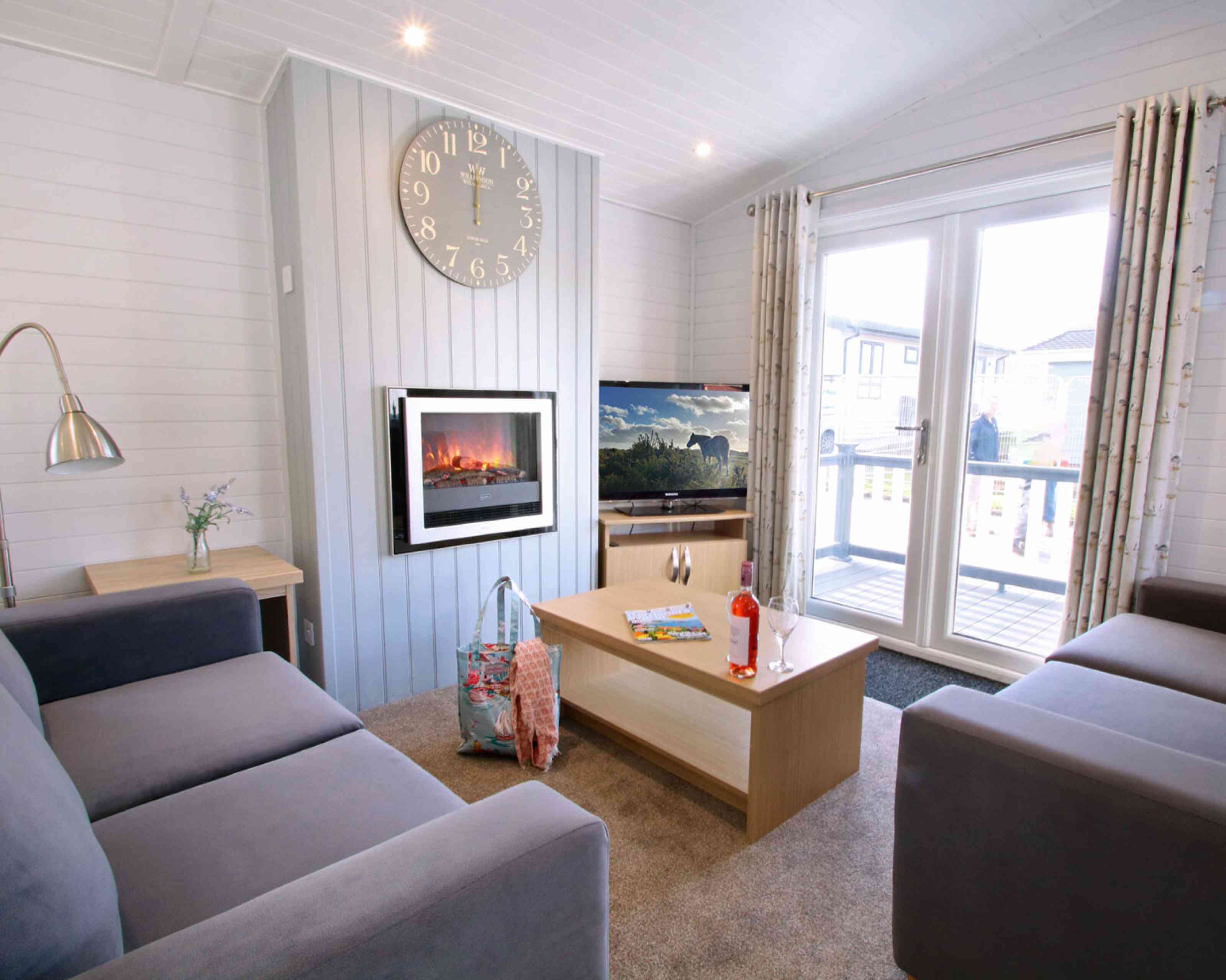 A cozy living room featuring two gray sofas, a coffee table with magazines and a drink, and a wall-mounted electric fireplace. A large clock hangs on the wall, and sliding glass doors open to a deck with outdoor views.