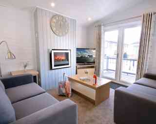 A cozy living room featuring two gray sofas, a coffee table with magazines and a drink, and a wall-mounted electric fireplace. A large clock hangs on the wall, and sliding glass doors open to a deck with outdoor views.