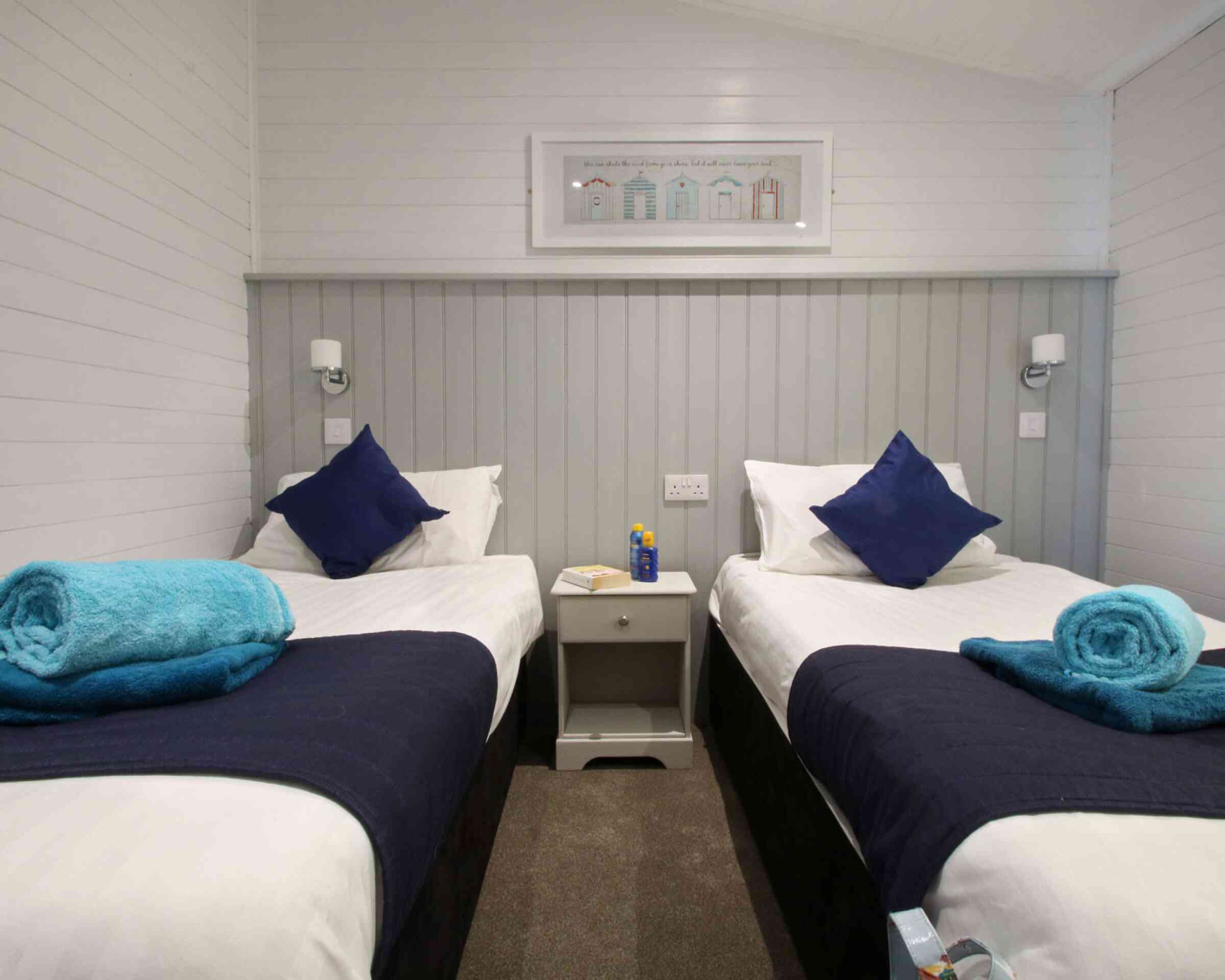 A cozy bedroom featuring two single beds with white linens and navy blue throws. Each bed has a blue pillow and a rolled blue towel on top. A small nightstand is placed between the beds, with a bottle and a notepad. The walls are white with a gray panel accent, and there's artwork of colorful houses above the beds.