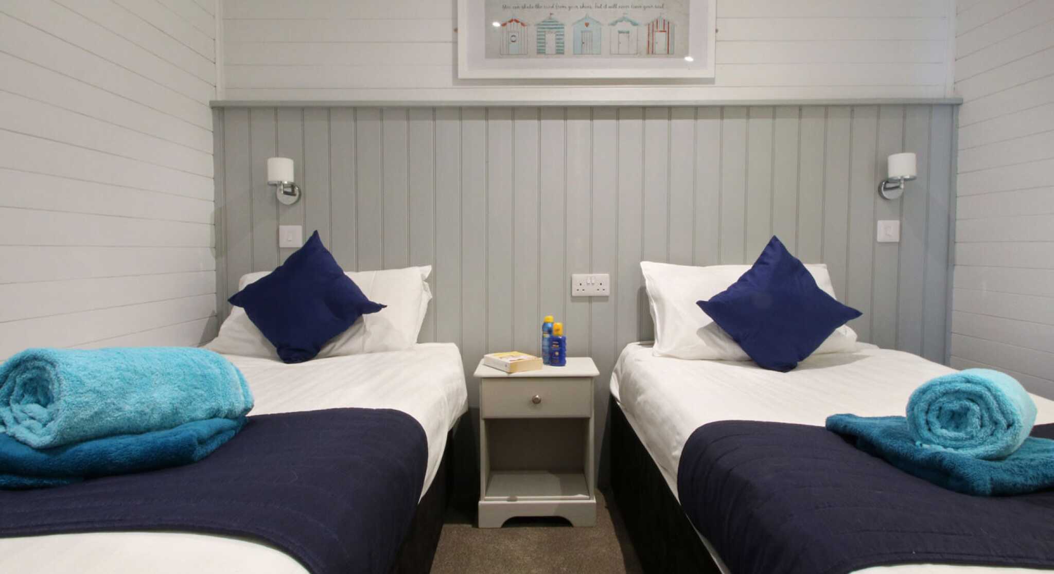 A cozy bedroom featuring two single beds with white linens and navy blue throws. Each bed has a blue pillow and a rolled blue towel on top. A small nightstand is placed between the beds, with a bottle and a notepad. The walls are white with a gray panel accent, and there's artwork of colorful houses above the beds.