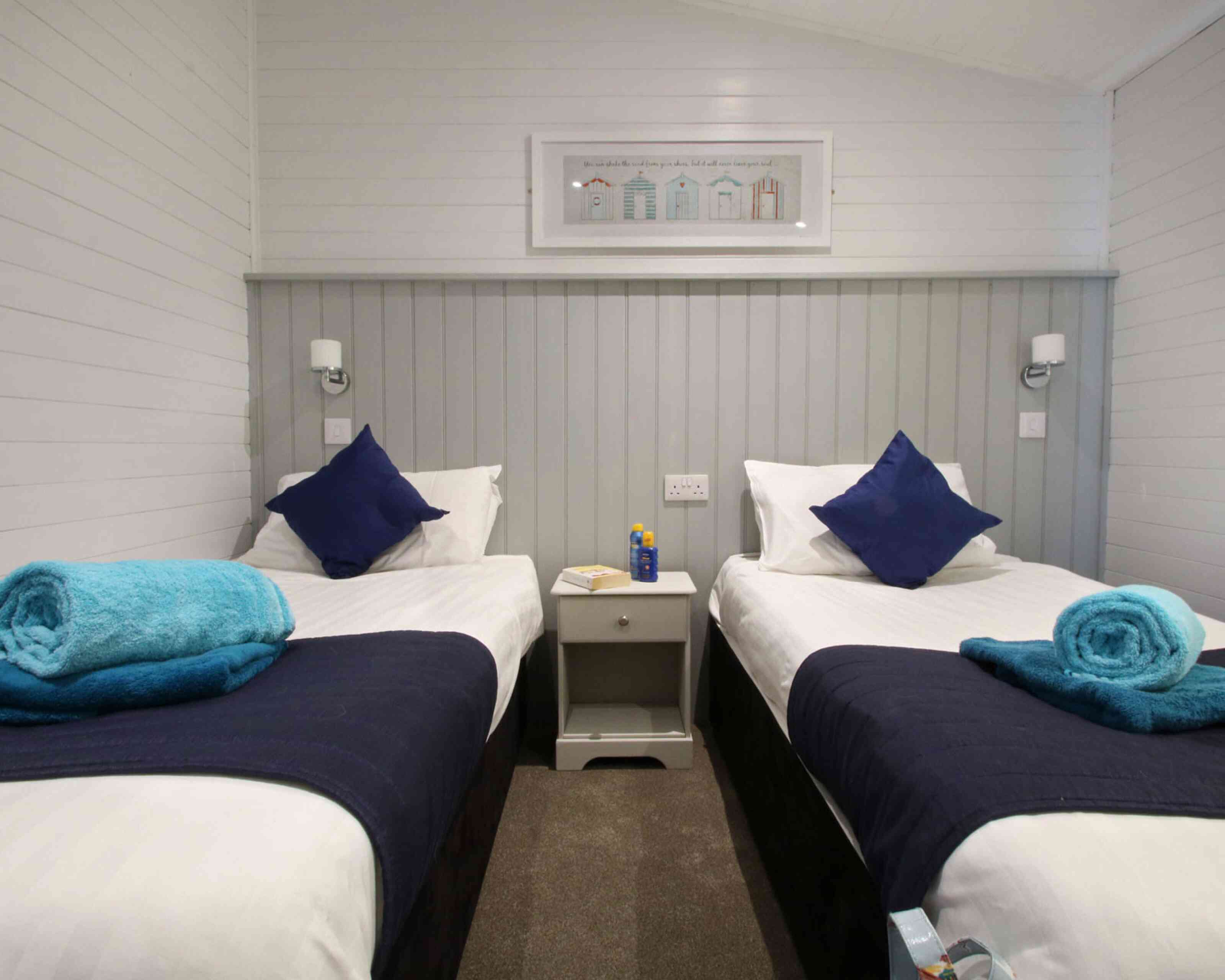A cozy bedroom featuring two single beds with white linens and navy blue throws. Each bed has a blue pillow and a rolled blue towel on top. A small nightstand is placed between the beds, with a bottle and a notepad. The walls are white with a gray panel accent, and there's artwork of colorful houses above the beds.