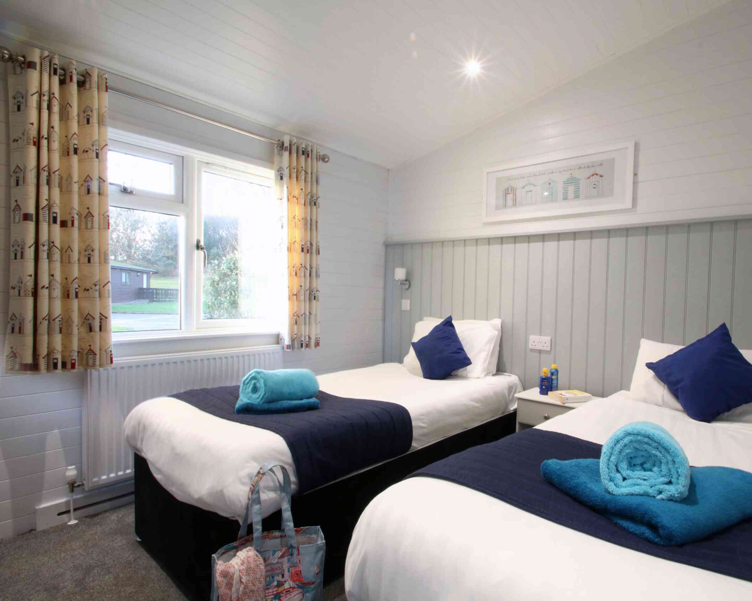 A cozy bedroom featuring two twin beds with white linens, dark blue bed runners, and bright blue towels. The room has a light gray wood-paneled wall, curtains adorned with a whimsical house pattern, and a window providing natural light. A small bedside table with a lamp and decorative art above the beds adds charm.