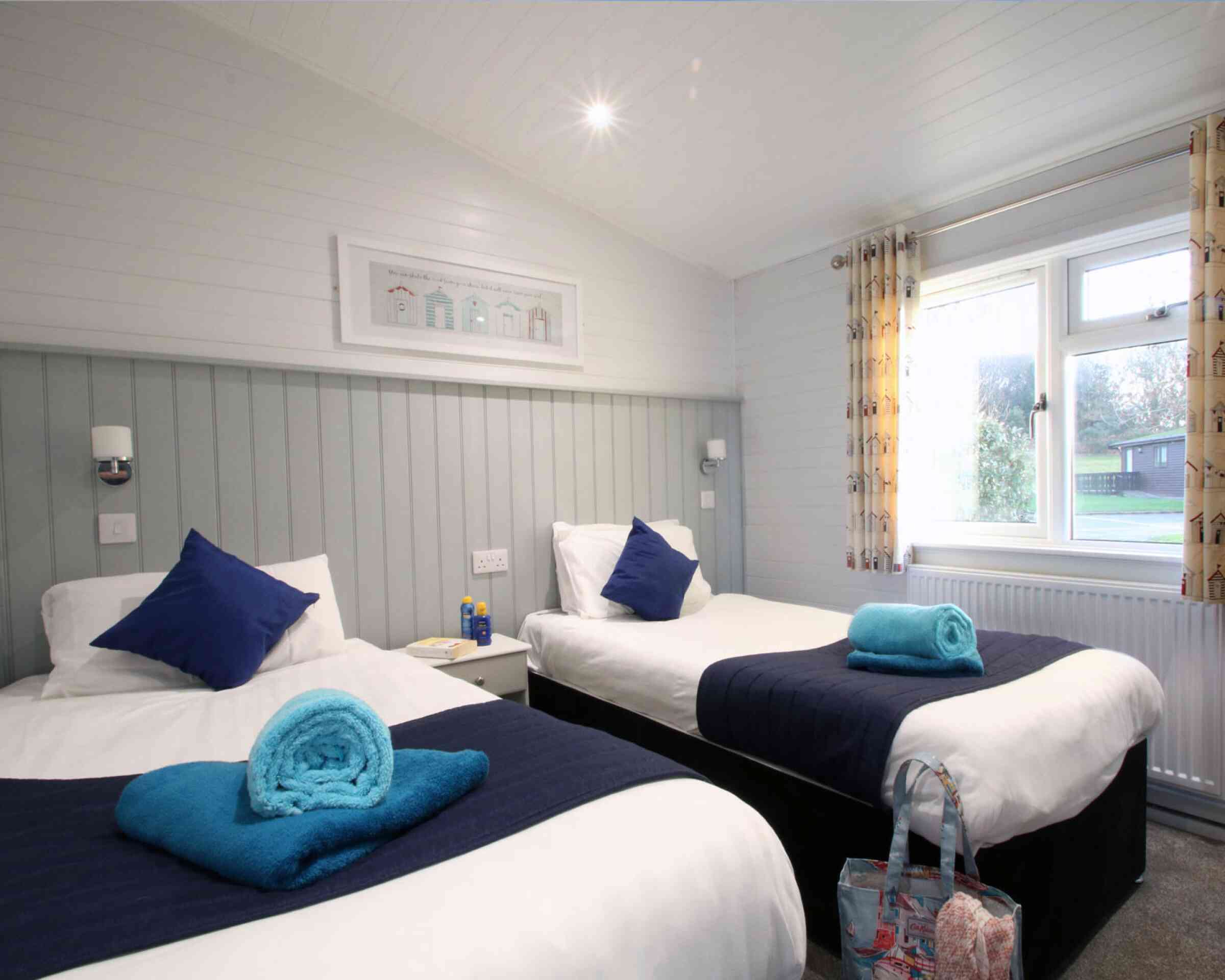 A cozy guest room featuring two single beds with blue throw pillows and towels. Light-colored walls and soft lighting create a welcoming atmosphere. A window with patterned curtains provides natural light, and a small bedside table holds a water bottle and other items.