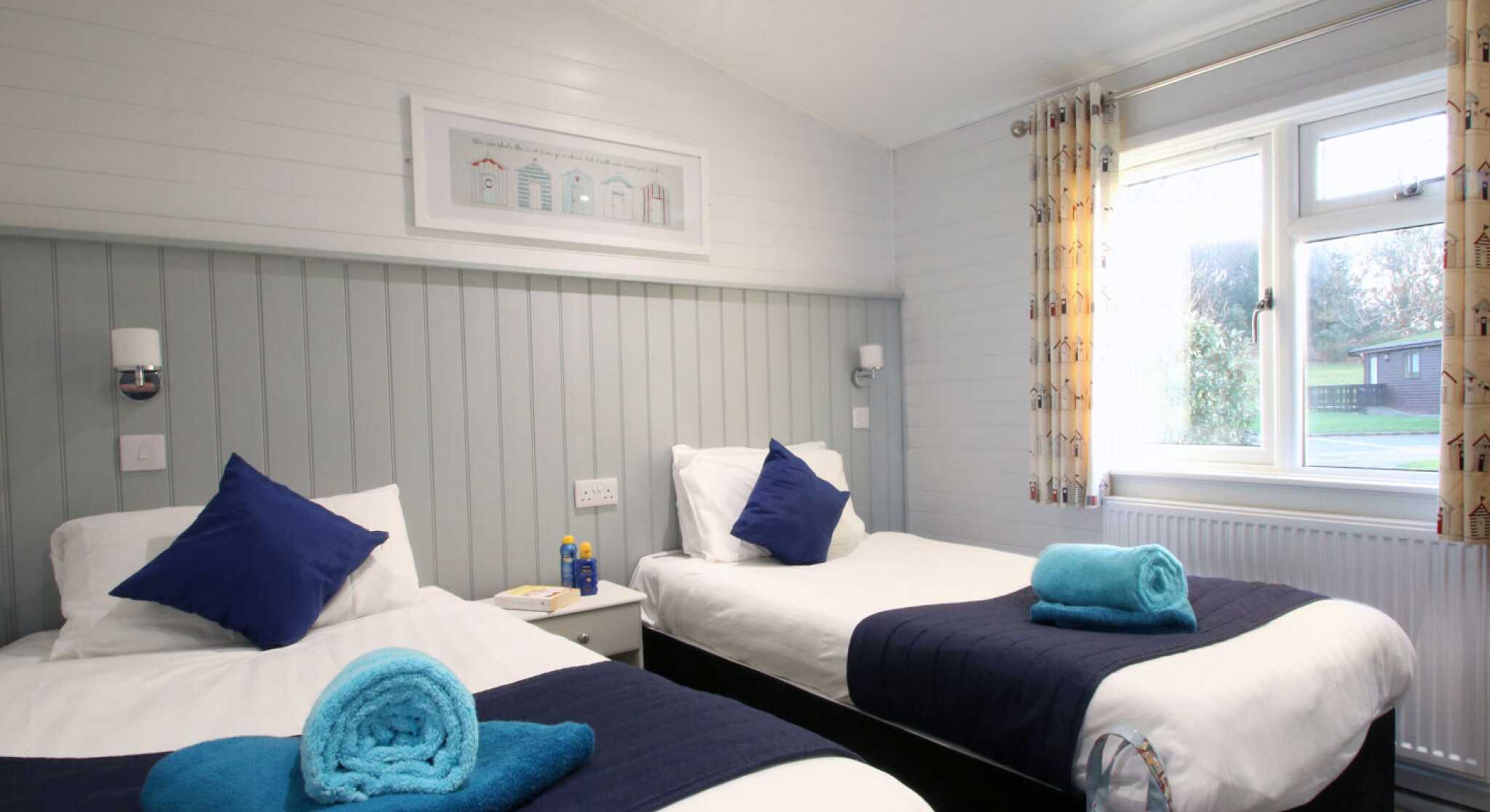 A cozy guest room featuring two single beds with blue throw pillows and towels. Light-colored walls and soft lighting create a welcoming atmosphere. A window with patterned curtains provides natural light, and a small bedside table holds a water bottle and other items.