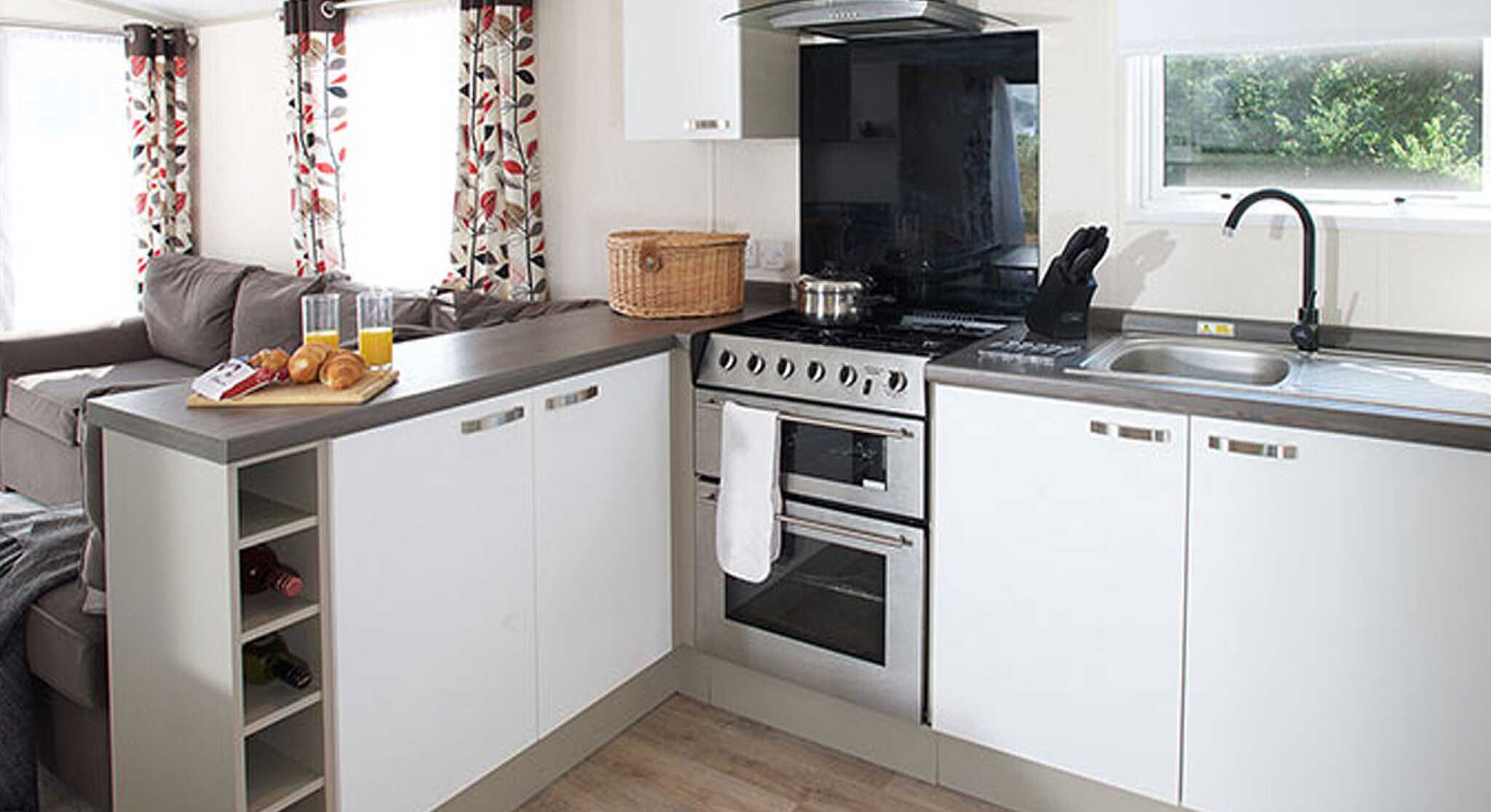 A modern kitchen with white cabinetry and a gray countertop. The kitchen features a stainless steel oven, a black extractor hood, and a sink with a dish rack. There is a wicker basket on the counter and a small wine rack integrated into the cabinetry. Natural light filters in through a window with white shades, and floral-patterned curtains frame the window. A cozy seating area with a gray sofa is visible in the background.