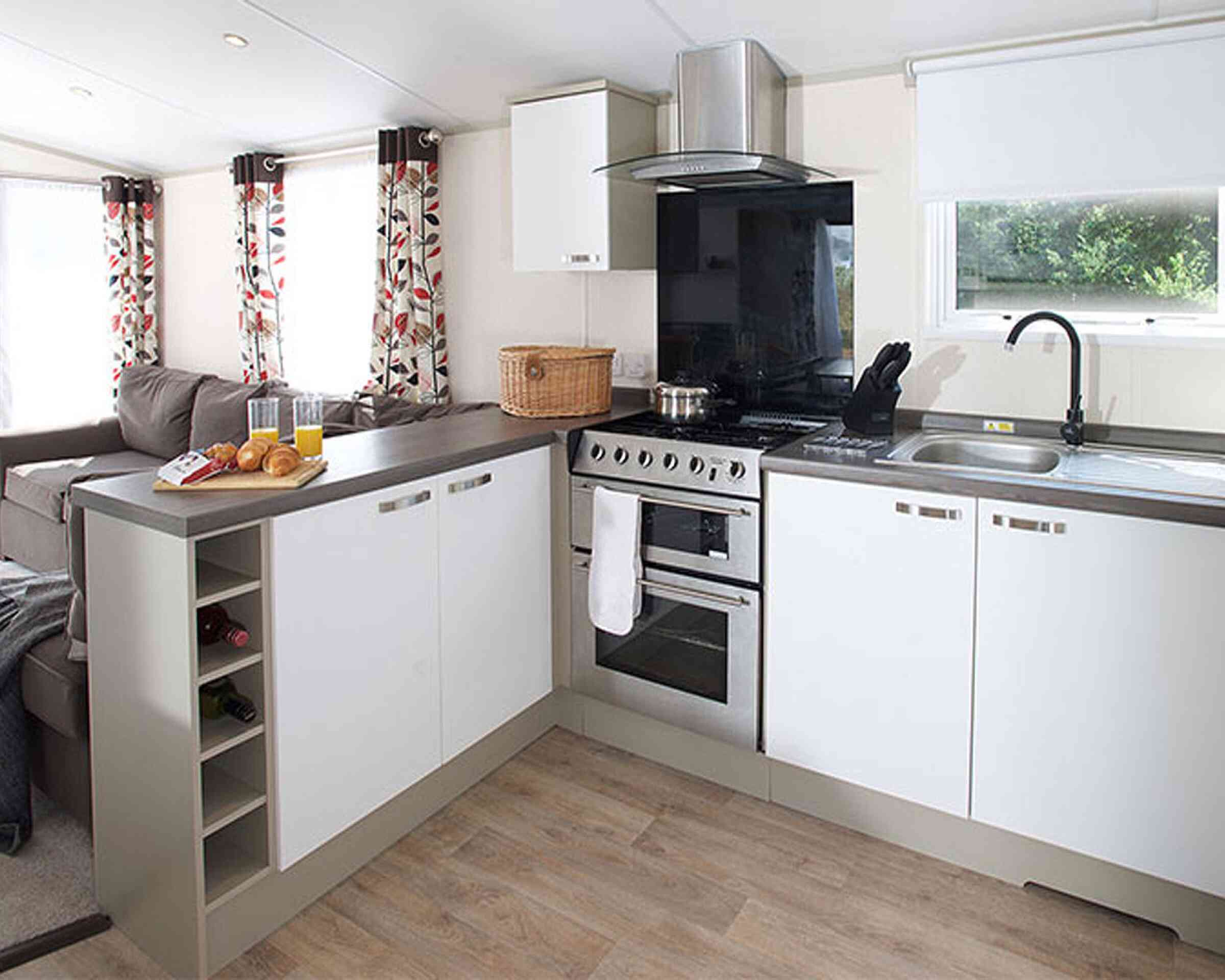 A modern kitchen with white cabinetry and a gray countertop. The kitchen features a stainless steel oven, a black extractor hood, and a sink with a dish rack. There is a wicker basket on the counter and a small wine rack integrated into the cabinetry. Natural light filters in through a window with white shades, and floral-patterned curtains frame the window. A cozy seating area with a gray sofa is visible in the background.