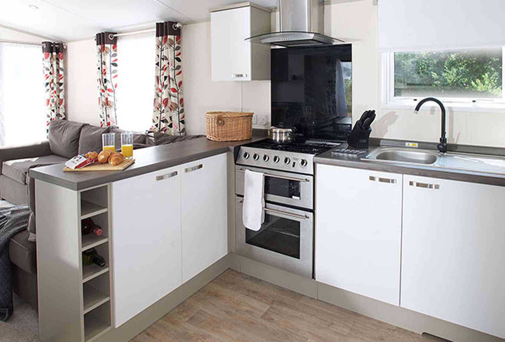 A modern kitchen with white cabinetry and a gray countertop. The kitchen features a stainless steel oven, a black extractor hood, and a sink with a dish rack. There is a wicker basket on the counter and a small wine rack integrated into the cabinetry. Natural light filters in through a window with white shades, and floral-patterned curtains frame the window. A cozy seating area with a gray sofa is visible in the background.