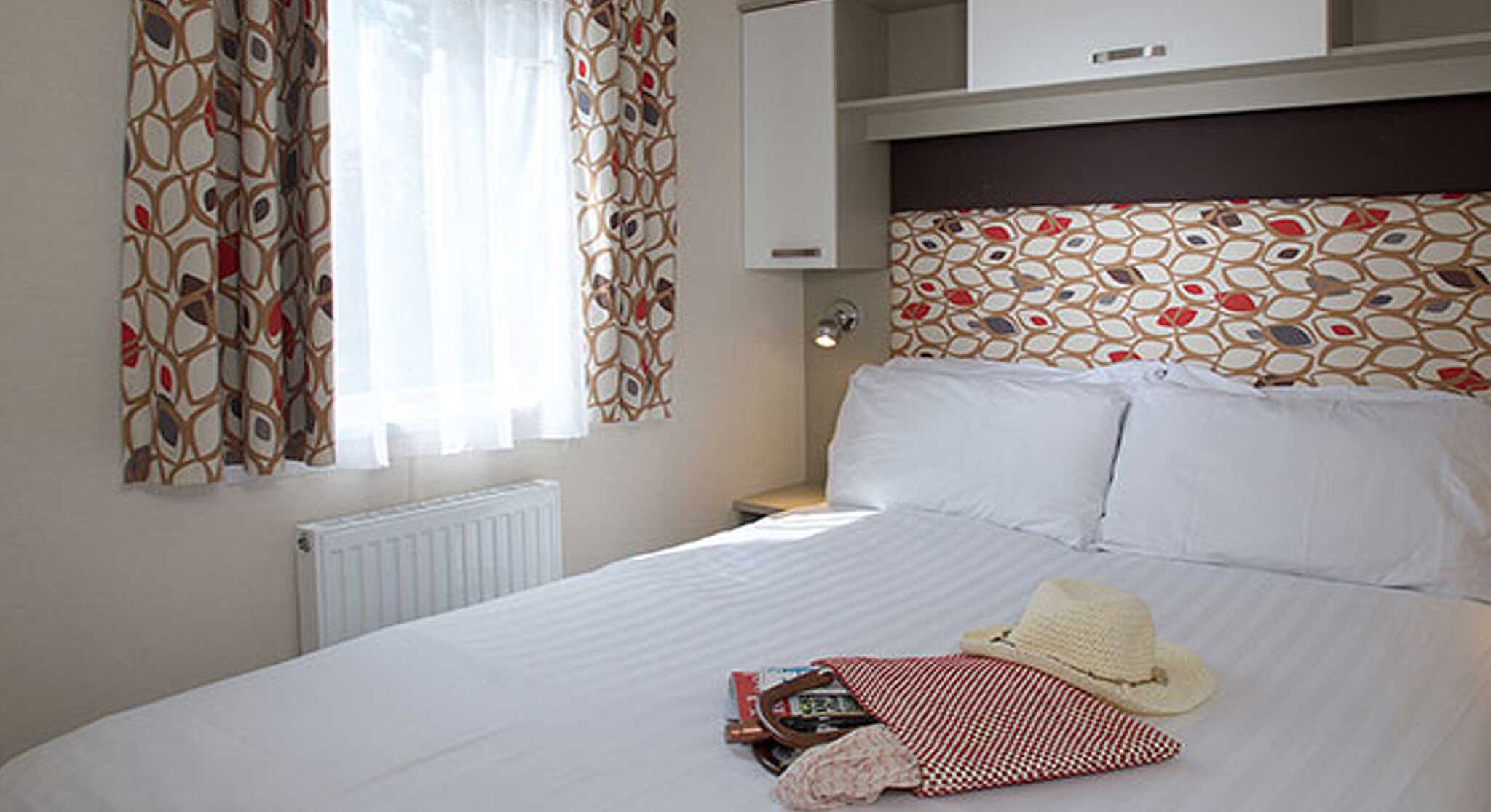 A cozy bedroom featuring a spacious double bed with crisp white linens. Colorful patterned curtains frame a window that lets in natural light. A decorative headboard adds a stylish touch, while a small bedside lamp and a few personal items like a hat and a small bag rest on the bed.