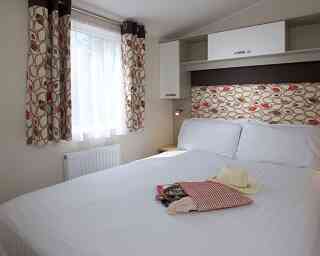 A cozy bedroom featuring a spacious double bed with crisp white linens. Colorful patterned curtains frame a window that lets in natural light. A decorative headboard adds a stylish touch, while a small bedside lamp and a few personal items like a hat and a small bag rest on the bed.