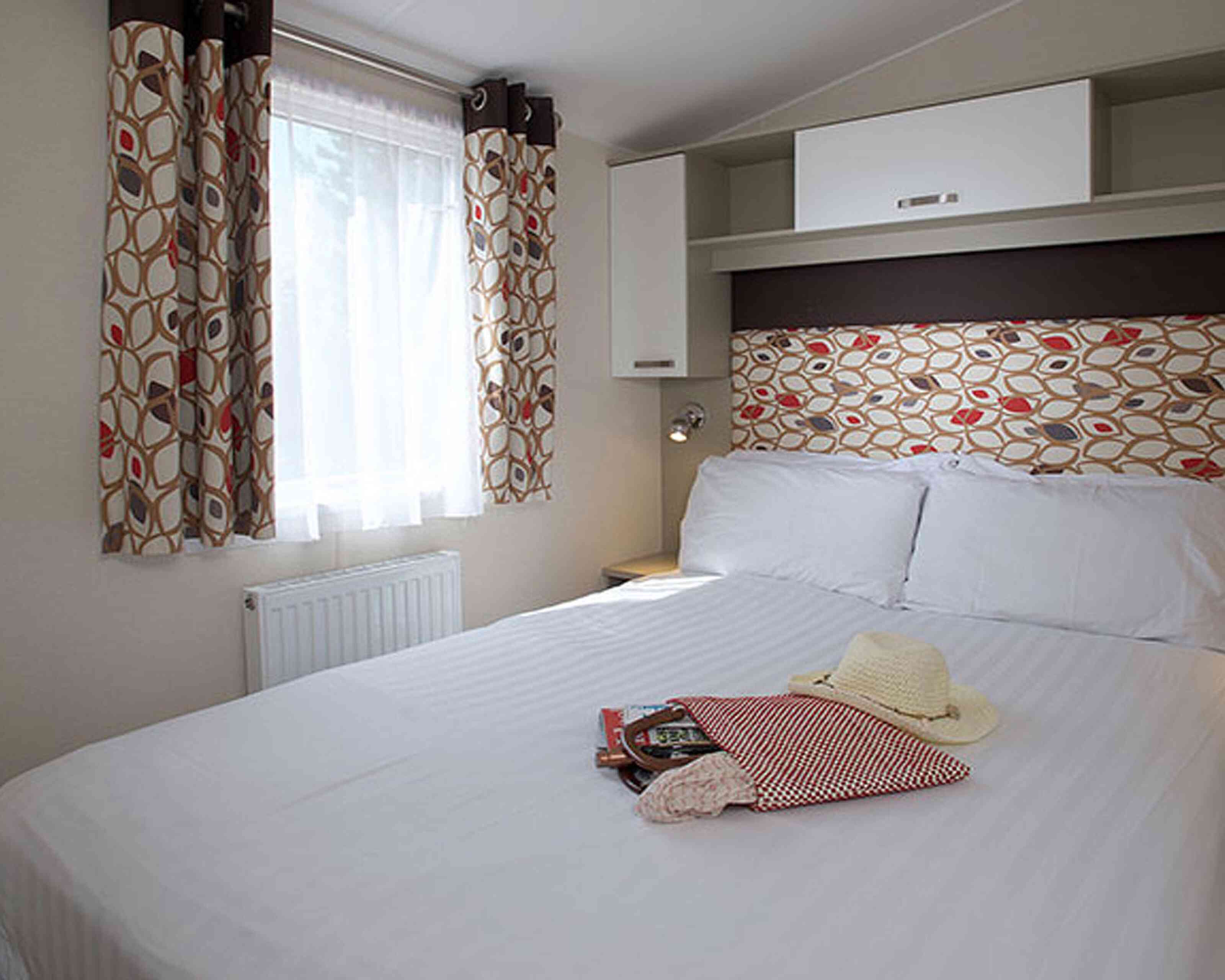 A cozy bedroom featuring a spacious double bed with crisp white linens. Colorful patterned curtains frame a window that lets in natural light. A decorative headboard adds a stylish touch, while a small bedside lamp and a few personal items like a hat and a small bag rest on the bed.