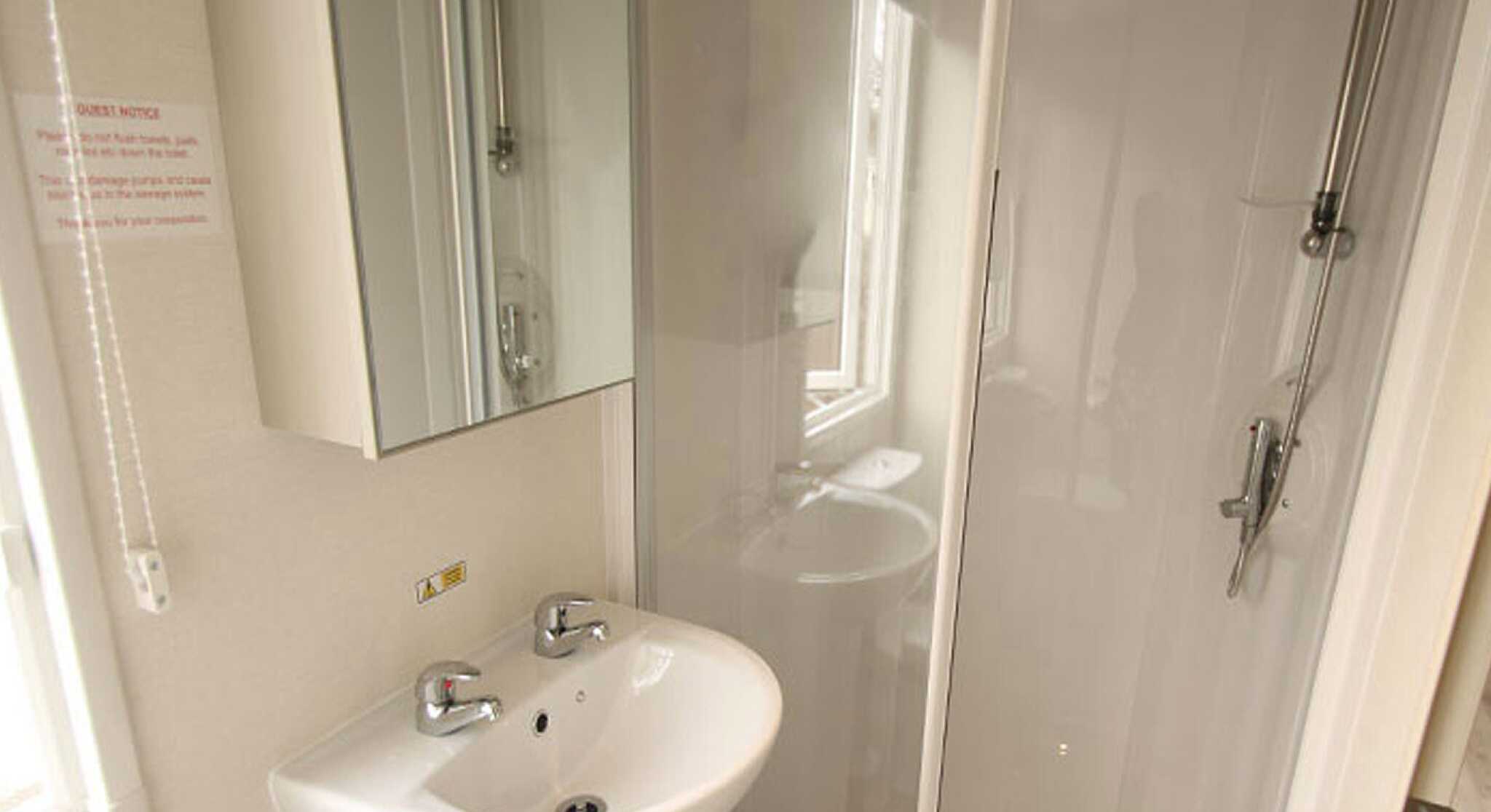 Bright bathroom featuring a small sink with two faucets, a shower stall with transparent glass doors, and a mirror mounted on the wall. Natural light enters through a window.