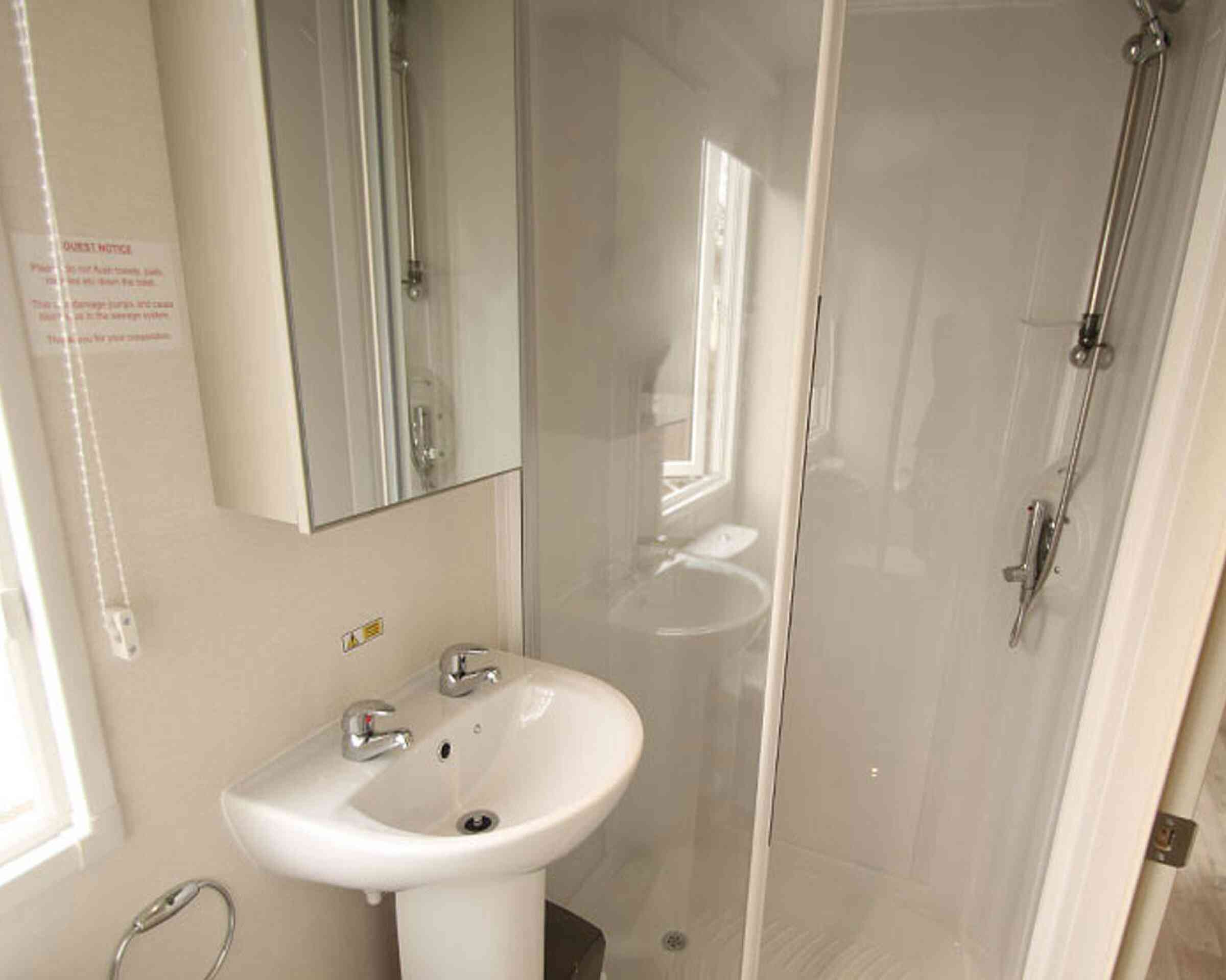 Bright bathroom featuring a small sink with two faucets, a shower stall with transparent glass doors, and a mirror mounted on the wall. Natural light enters through a window.