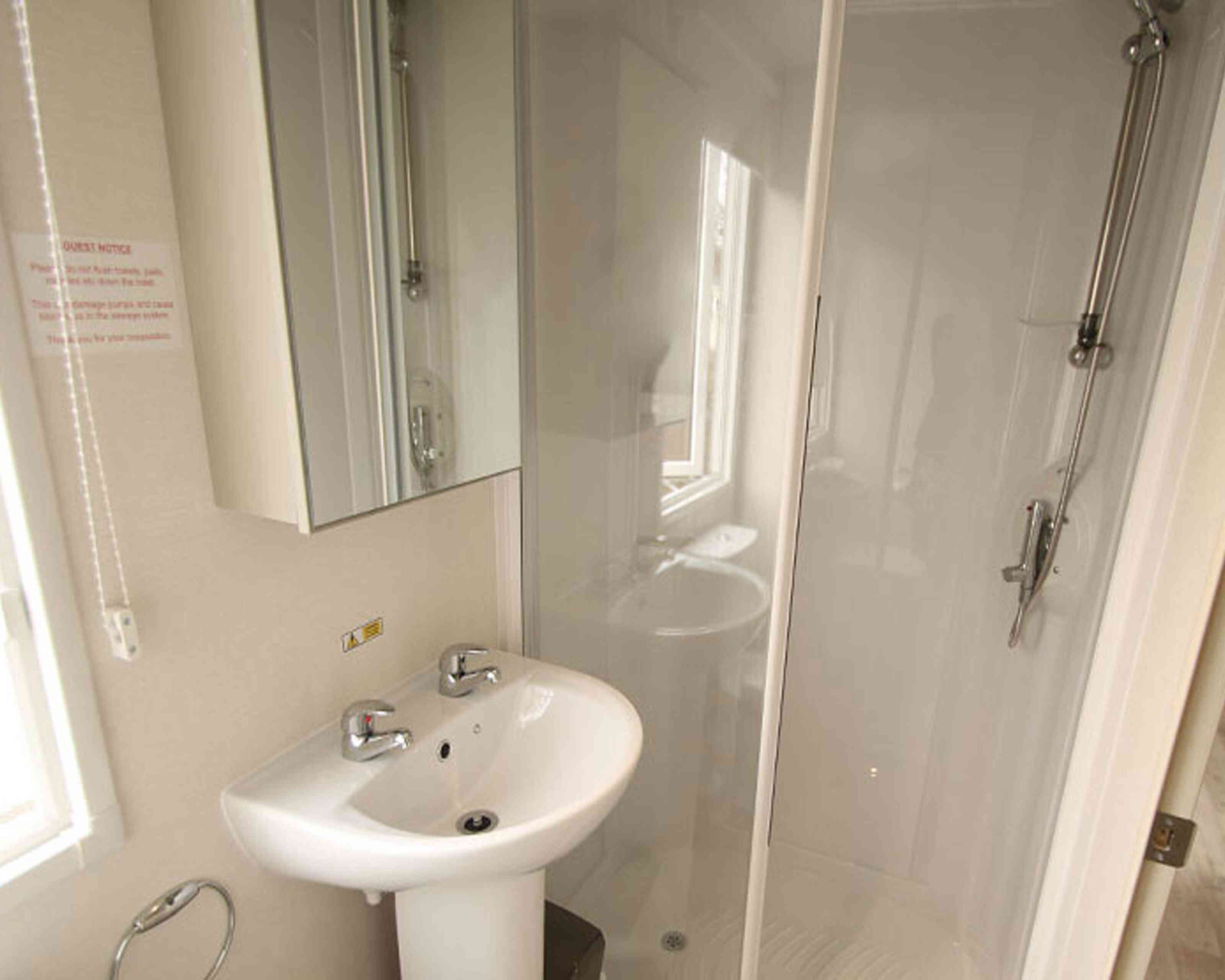 Bright bathroom featuring a small sink with two faucets, a shower stall with transparent glass doors, and a mirror mounted on the wall. Natural light enters through a window.
