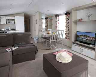 Cozy interior of a modern mobile home with a brown sectional sofa, a coffee table, and a dining area with white chairs. Natural light streams in through the large windows decorated with floral curtains. A television is mounted on a shelf, and a kitchen area is visible in the background.