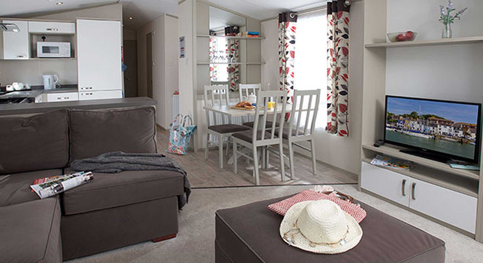 Cozy interior of a modern mobile home with a brown sectional sofa, a coffee table, and a dining area with white chairs. Natural light streams in through the large windows decorated with floral curtains. A television is mounted on a shelf, and a kitchen area is visible in the background.