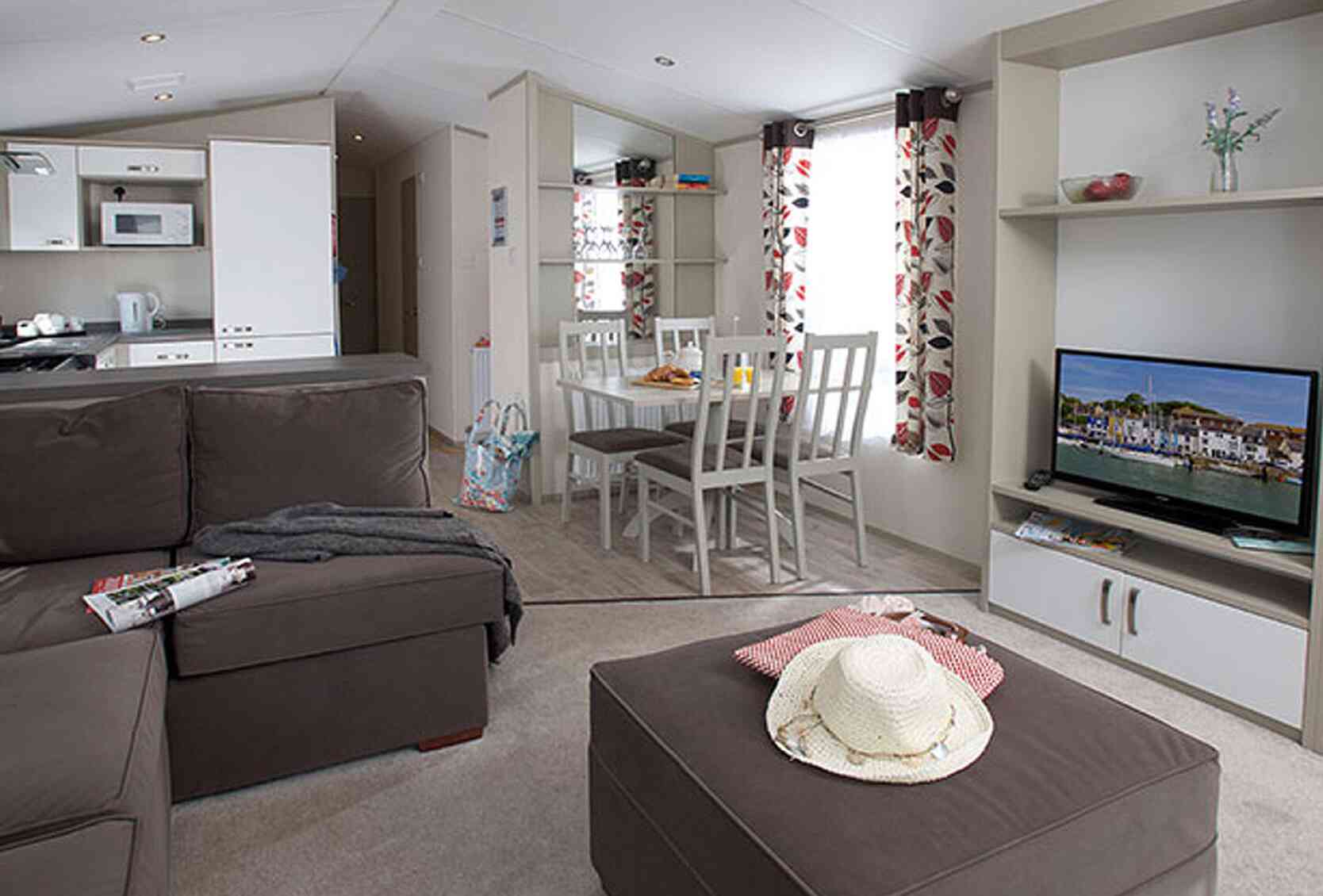 Cozy interior of a modern mobile home with a brown sectional sofa, a coffee table, and a dining area with white chairs. Natural light streams in through the large windows decorated with floral curtains. A television is mounted on a shelf, and a kitchen area is visible in the background.