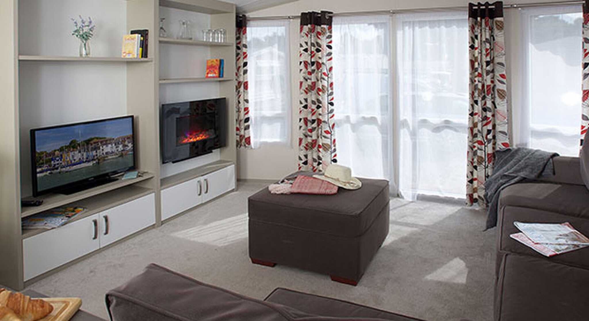 Cozy living room featuring a comfortable sofa, an ottoman, and a modern television. Bright curtains adorn the windows, allowing natural light in. A small TV unit with decorative items and a fireplace adds warmth to the space.