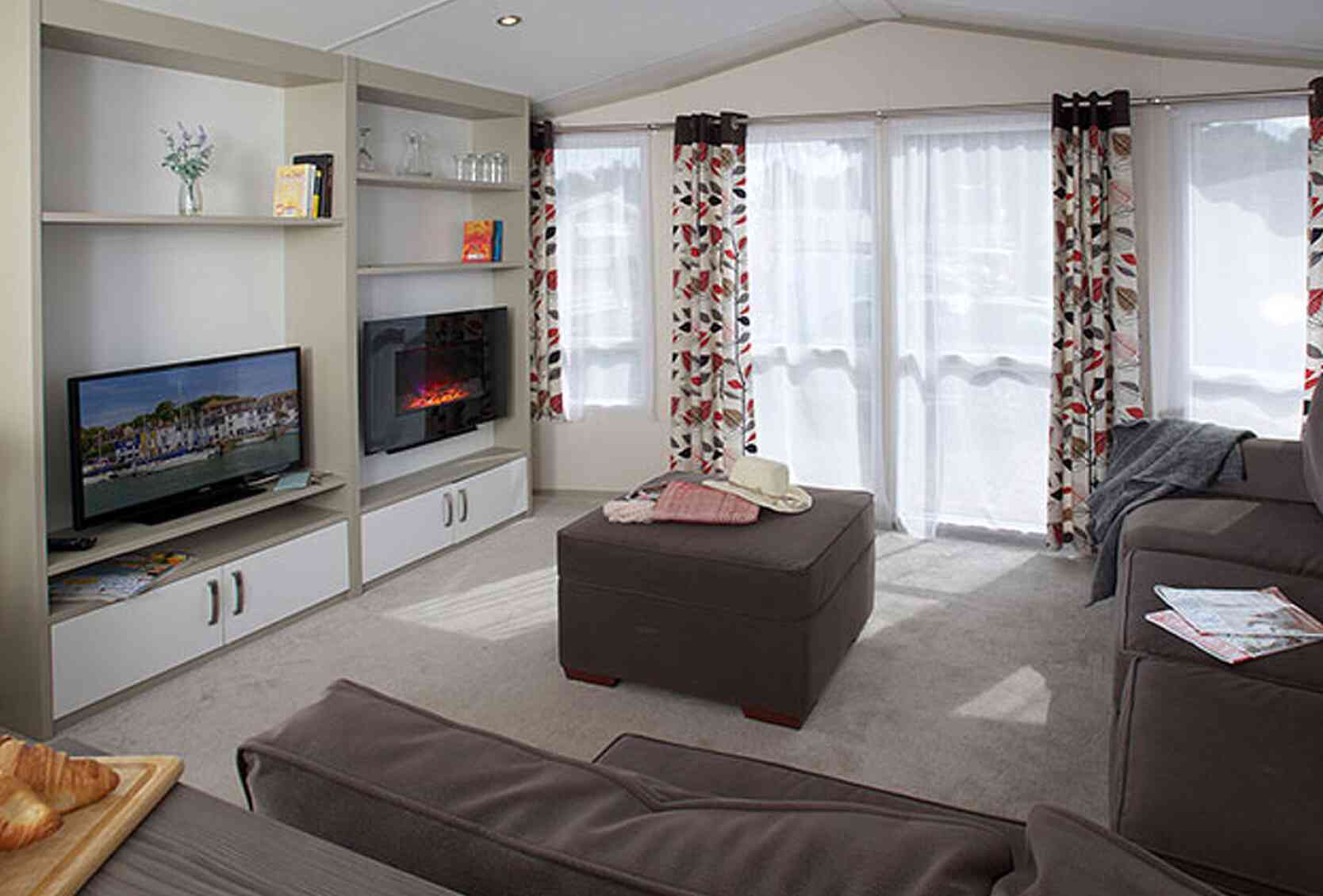 Cozy living room featuring a comfortable sofa, an ottoman, and a modern television. Bright curtains adorn the windows, allowing natural light in. A small TV unit with decorative items and a fireplace adds warmth to the space.