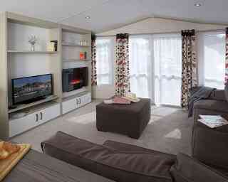Cozy living room featuring a comfortable sofa, an ottoman, and a modern television. Bright curtains adorn the windows, allowing natural light in. A small TV unit with decorative items and a fireplace adds warmth to the space.