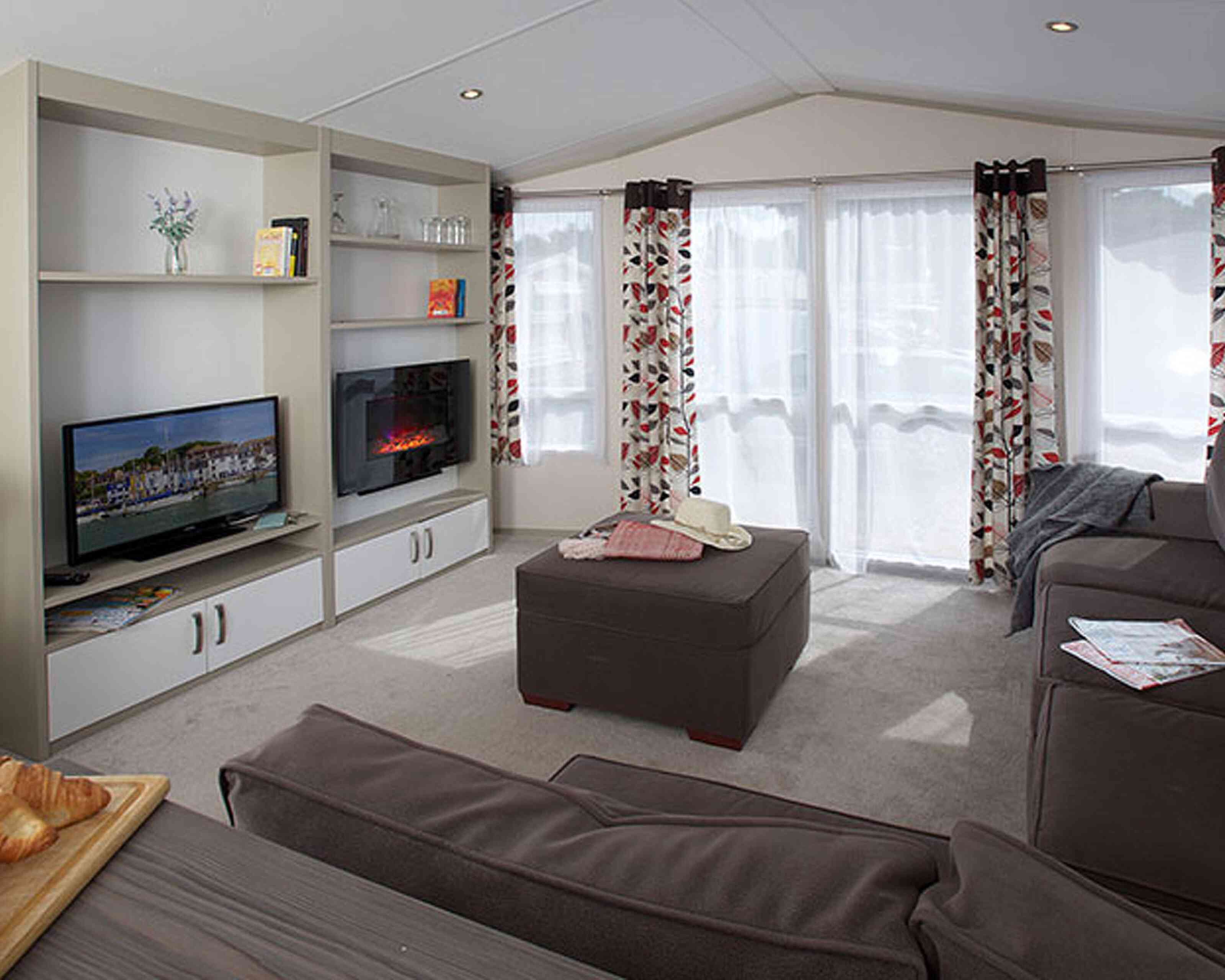 Cozy living room featuring a comfortable sofa, an ottoman, and a modern television. Bright curtains adorn the windows, allowing natural light in. A small TV unit with decorative items and a fireplace adds warmth to the space.