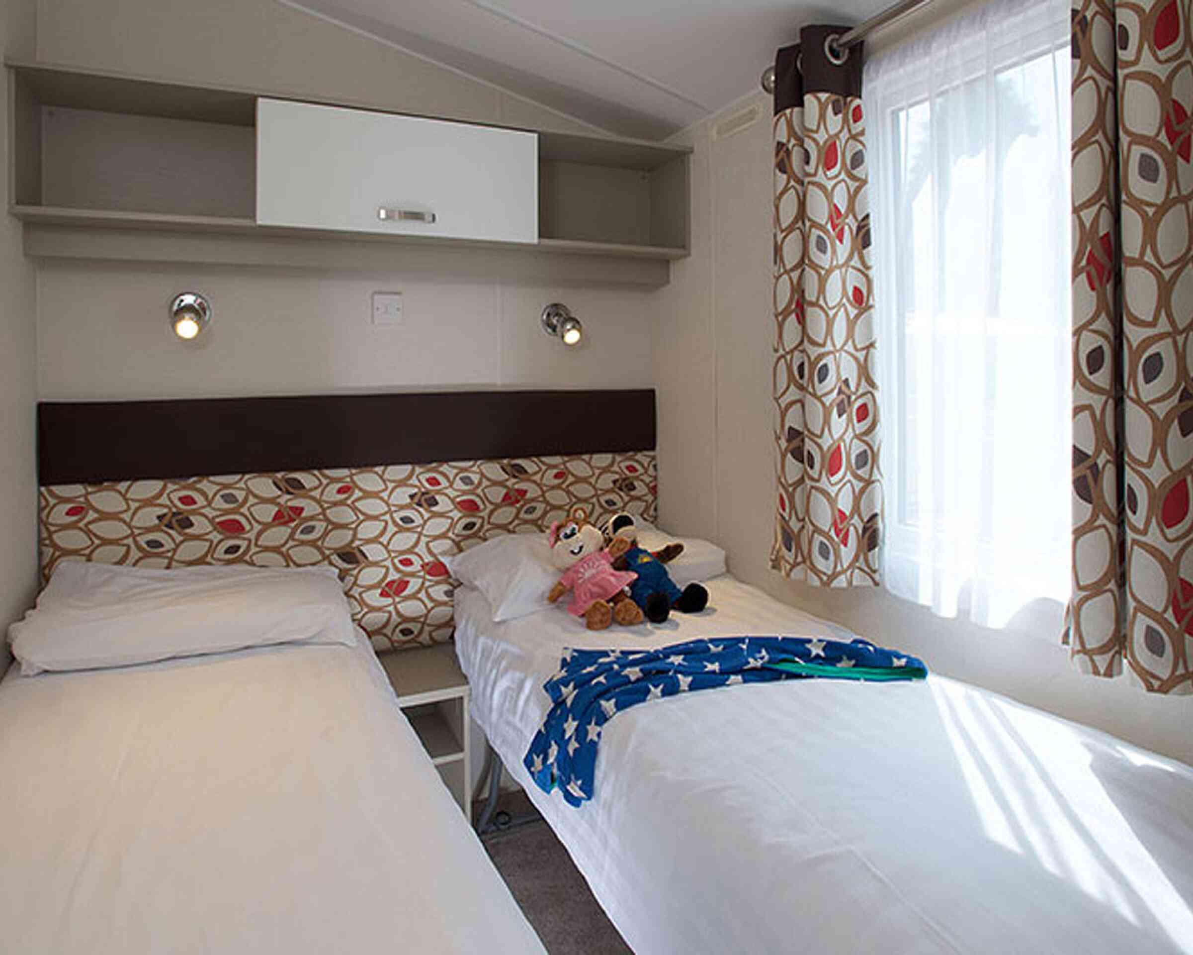 Two single beds with white linens positioned in a cozy room. A colorful patterned headboard separates the beds, and a plush toy sits on one. Bright curtains frame a window, and a shelf above provides storage.