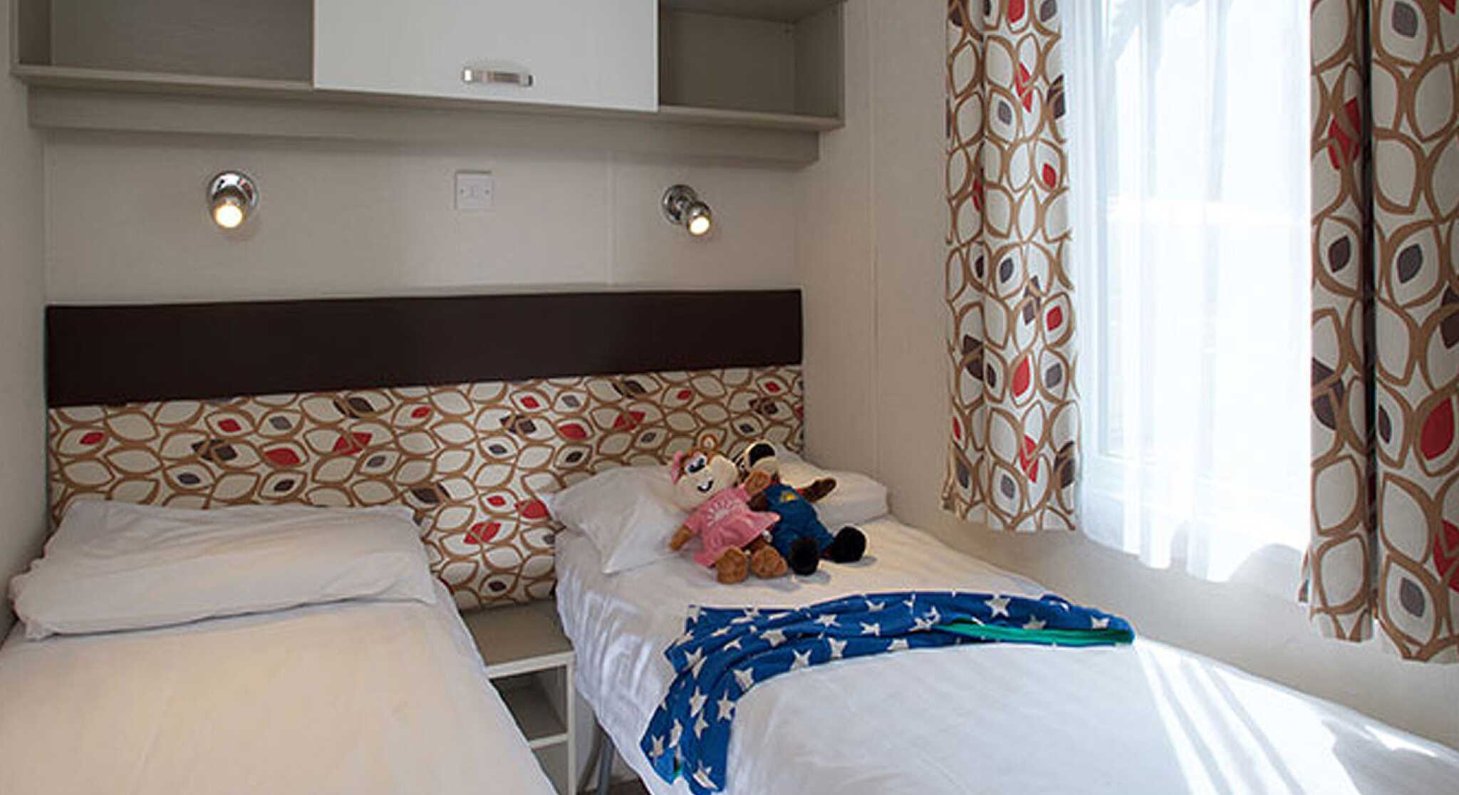 Two single beds with white linens positioned in a cozy room. A colorful patterned headboard separates the beds, and a plush toy sits on one. Bright curtains frame a window, and a shelf above provides storage.
