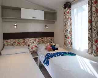 Two single beds with white linens positioned in a cozy room. A colorful patterned headboard separates the beds, and a plush toy sits on one. Bright curtains frame a window, and a shelf above provides storage.