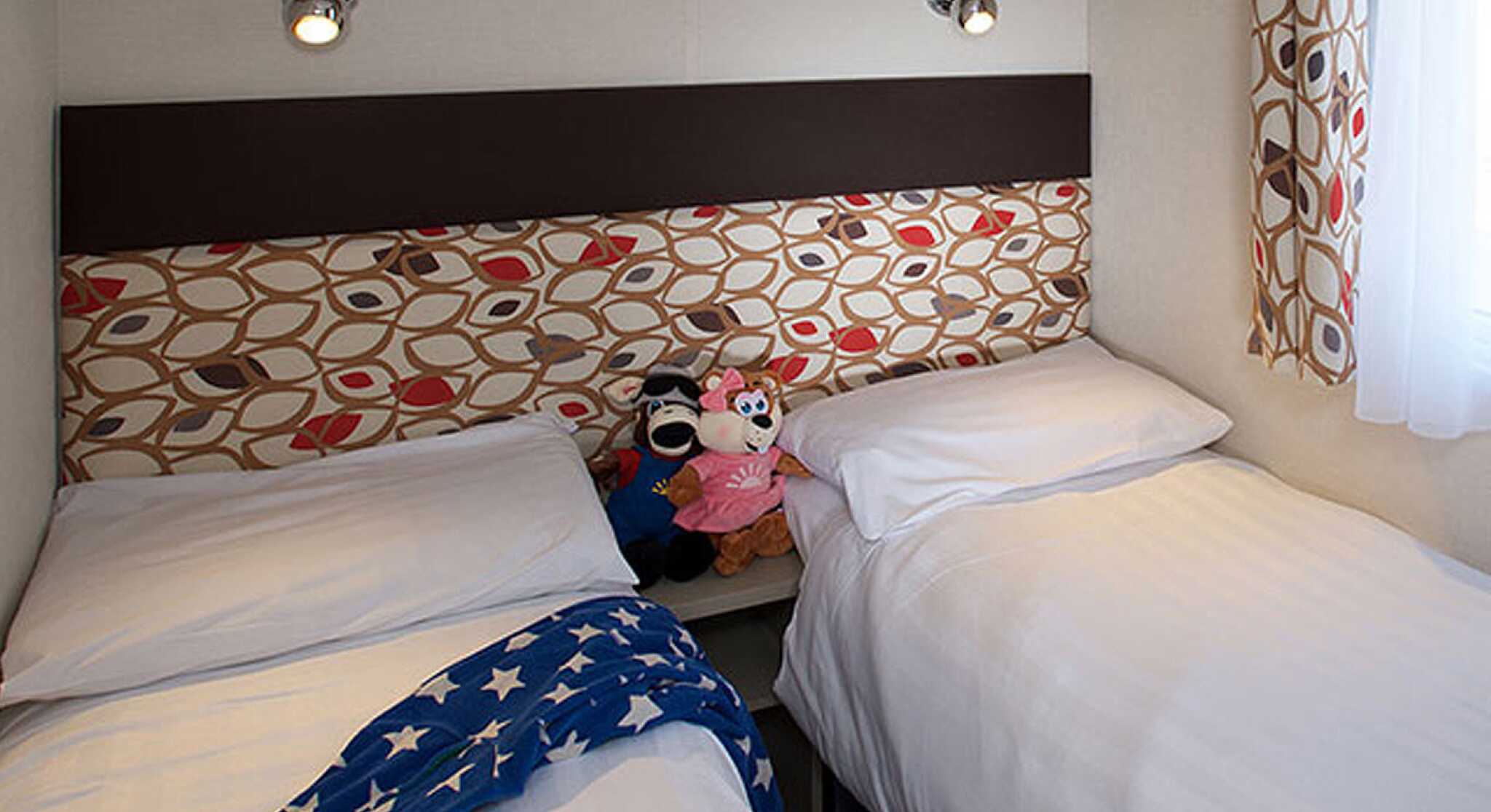 A cozy bedroom with two single beds, each covered with white linens. A colorful headboard features a pattern of red and brown leaves. A pair of plush toys, including a mouse and a pink doll, sit on the edge of one bed. A blue starry blanket is draped over the other bed. Soft lighting shines from wall-mounted fixtures.