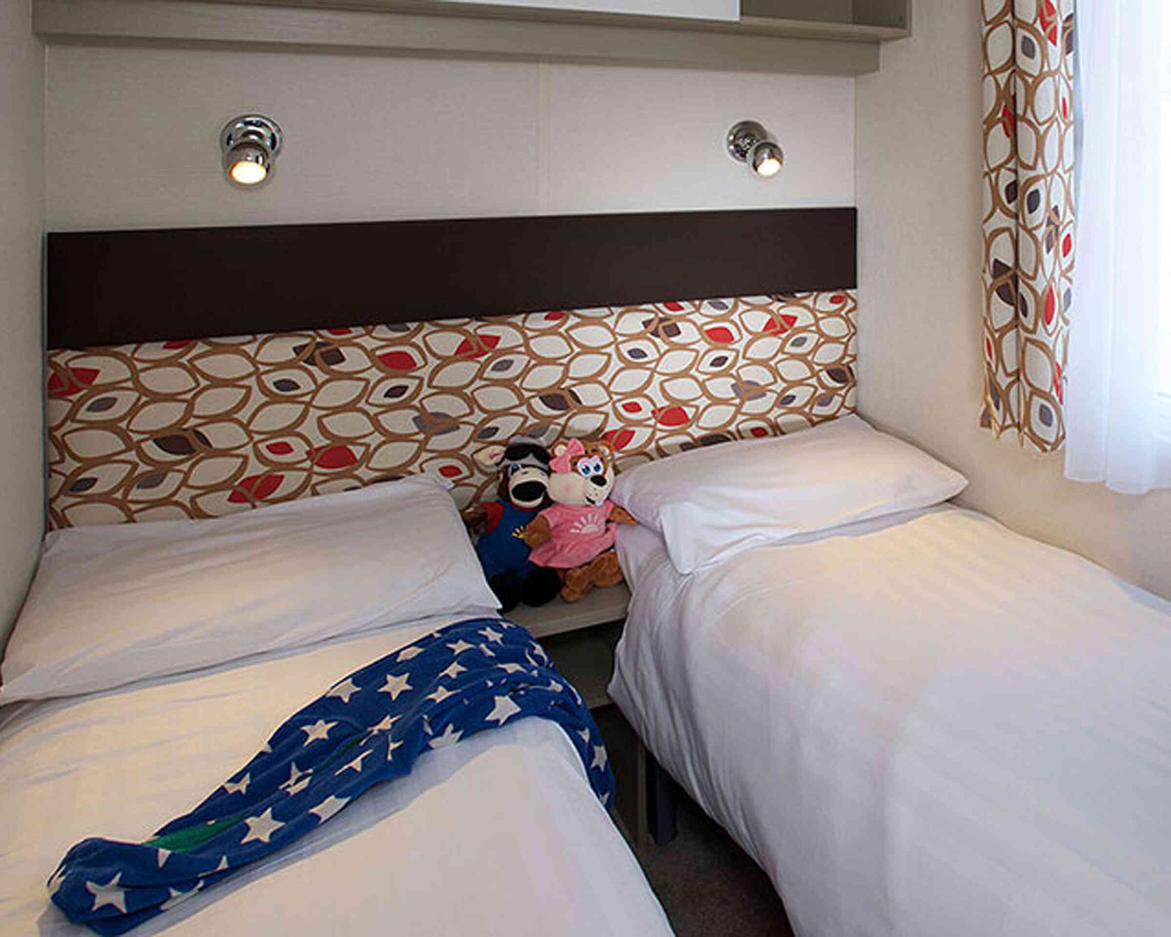 A cozy bedroom with two single beds, each covered with white linens. A colorful headboard features a pattern of red and brown leaves. A pair of plush toys, including a mouse and a pink doll, sit on the edge of one bed. A blue starry blanket is draped over the other bed. Soft lighting shines from wall-mounted fixtures.