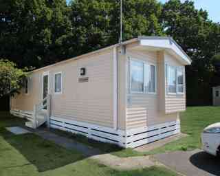 A light-colored mobile home with a small porch and steps, situated on a grassy area. Trees are visible in the background.