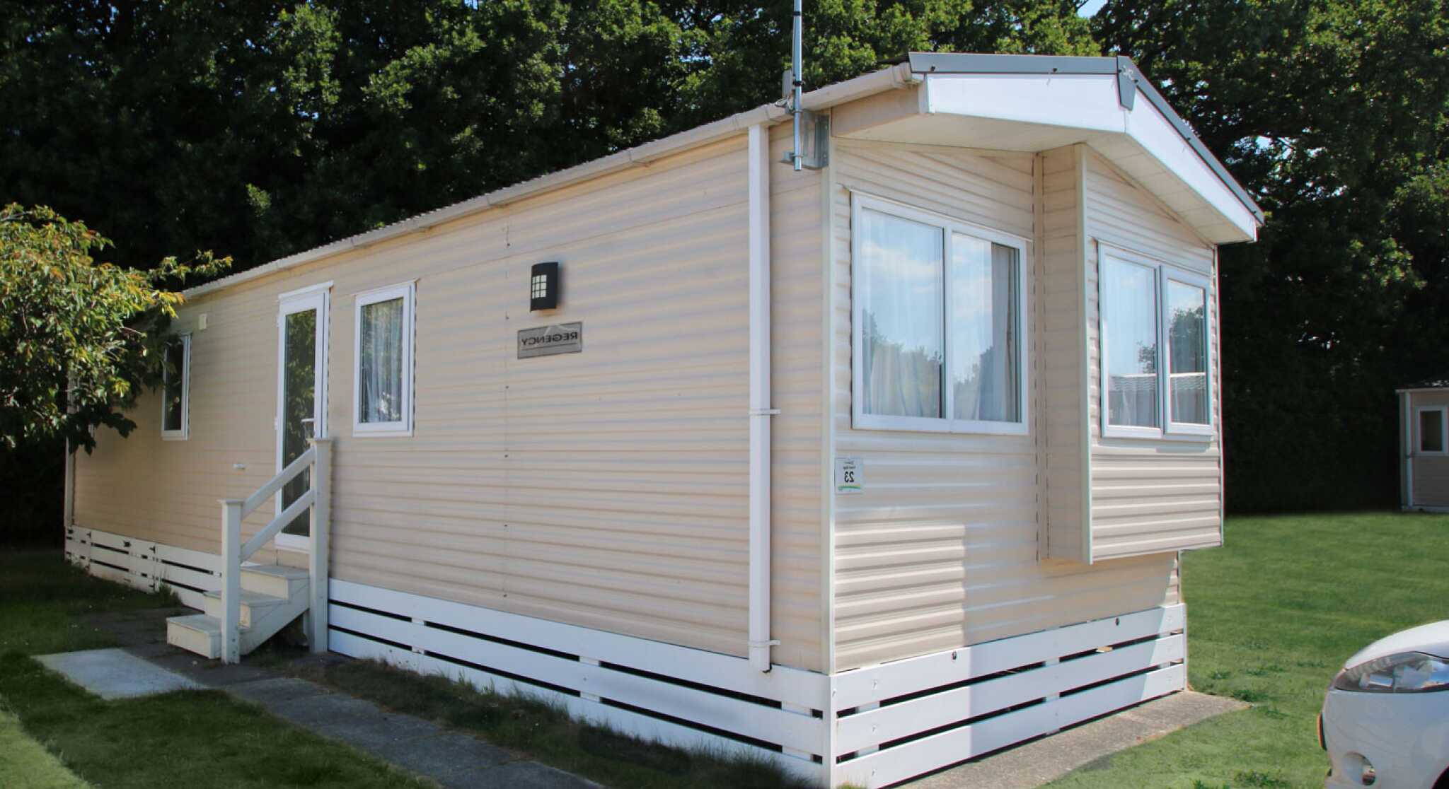 A light-colored mobile home with a small porch and steps, situated on a grassy area. Trees are visible in the background.