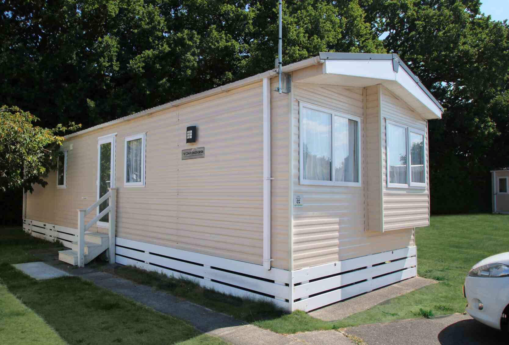 A light-colored mobile home with a small porch and steps, situated on a grassy area. Trees are visible in the background.