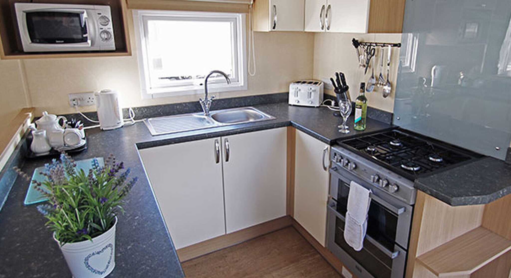 Modern kitchen featuring a sink, gas stove, microwave, and a toaster. Cabinets are made of light wood and white surfaces, with a countertop showcasing utensils and a small potted plant. The area is well-lit by natural light coming from a window.
