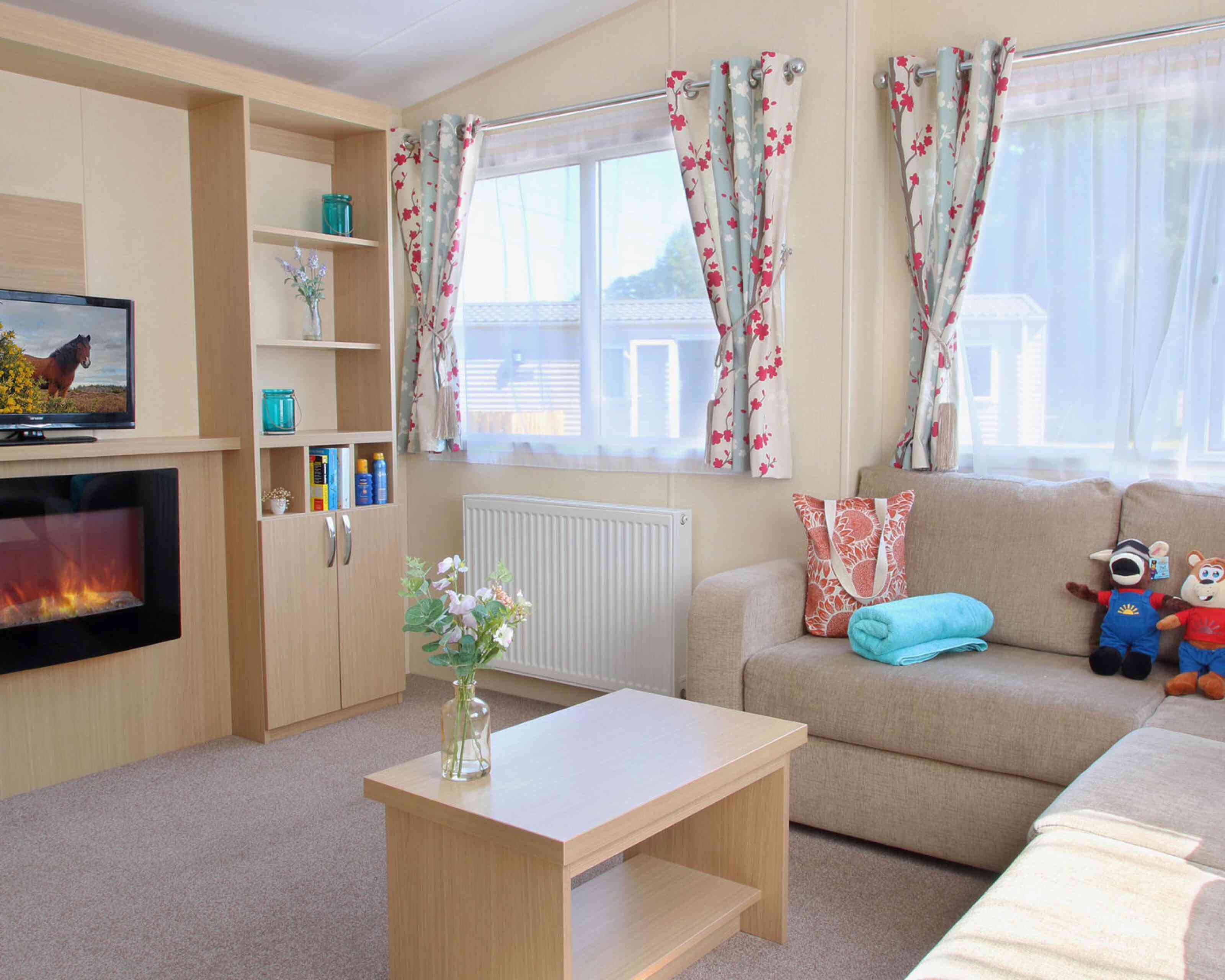 A cozy living room featuring a beige sofa, a wooden coffee table, and a decorative vase with flowers. There are colorful curtains with floral patterns, a television on a shelf, and a faux fireplace below it. A blue towel and stuffed toys are also on the sofa.